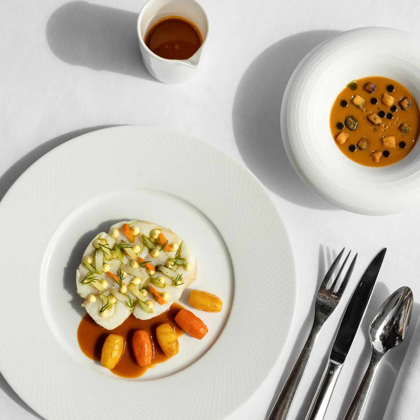 White plate with a culinary preparation based on fish, vegetables, and a sauce, accompanied by a bowl of soup and a small sauce container, cutlery next to it.