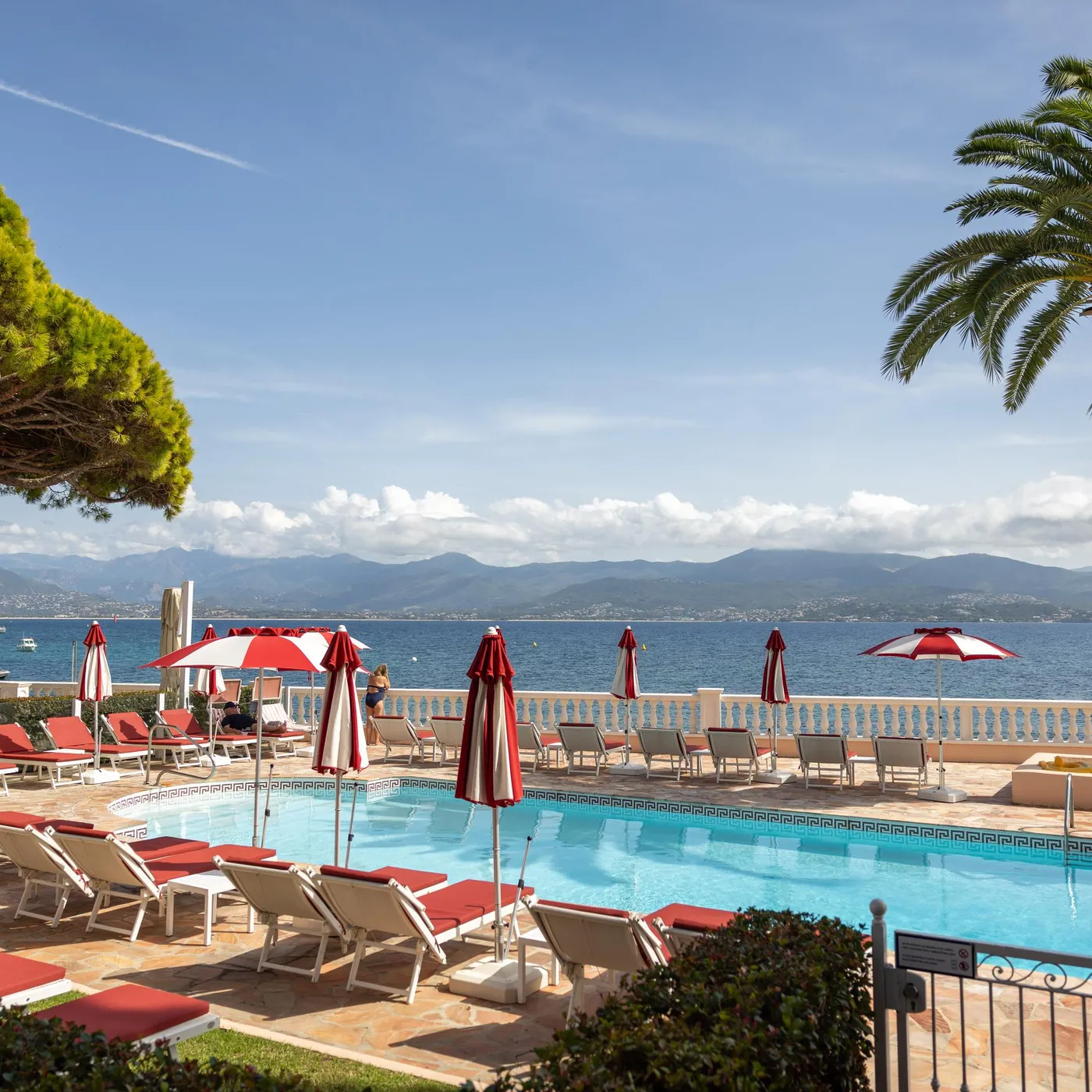 Piscine avec transats et parasols rouges et blancs, bordée de palmiers, face à une mer bleue avec des montagnes à l'horizon