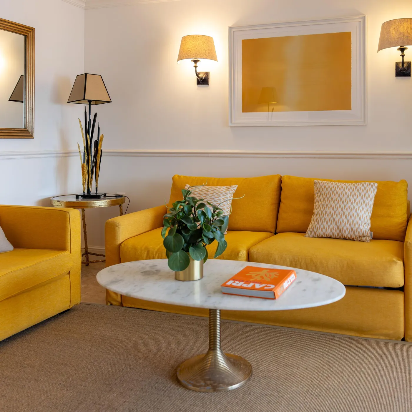 A living room with a sofa and a yellow armchair, a coffee table with a plant and a book, wall lamps, and empty frames on the walls.