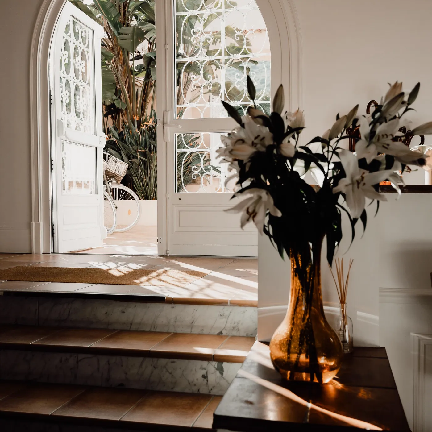 A vase with white flowers is placed on a console near an open glass door letting in the light.