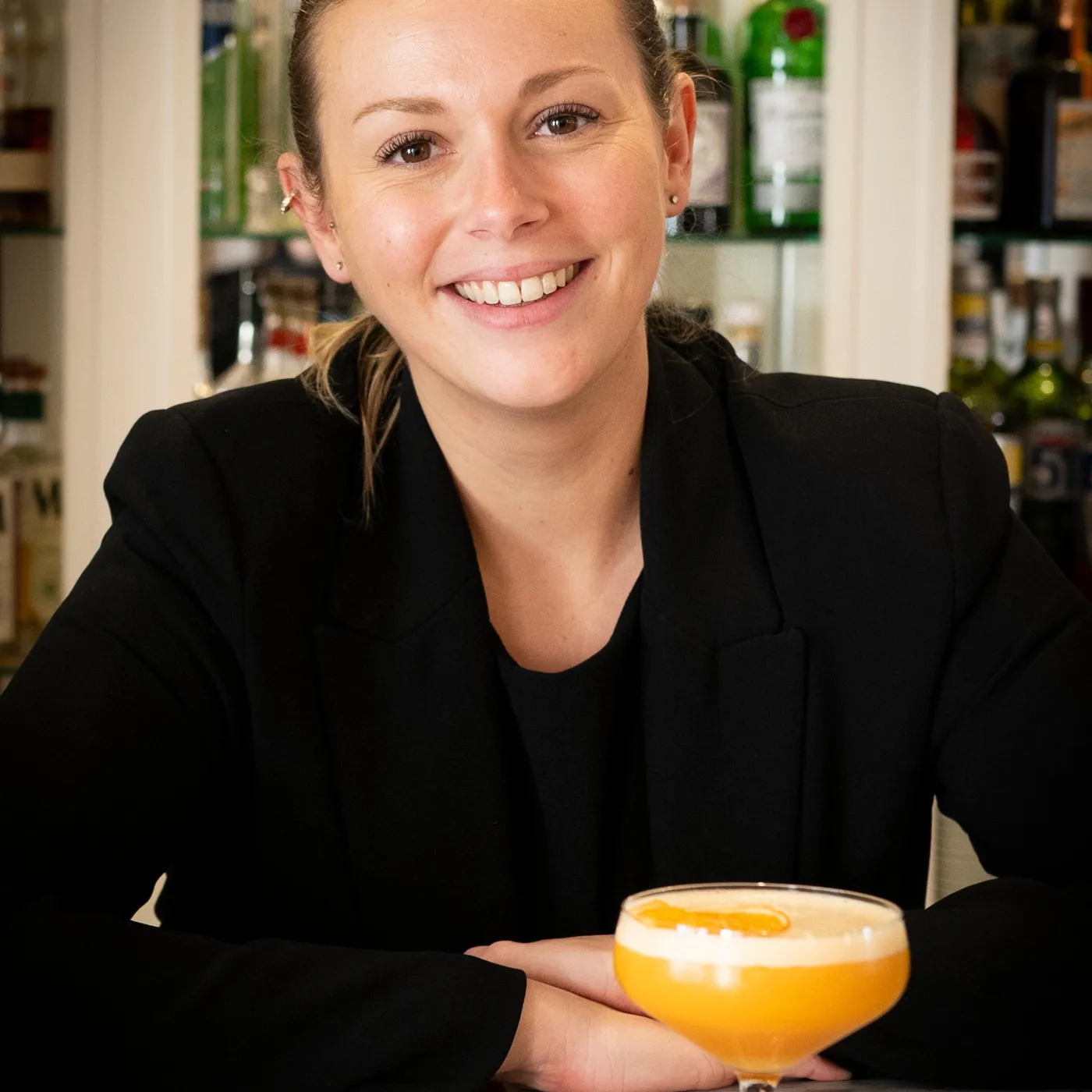 A smiling woman in black attire behind a bar holds a glass containing an orange cocktail.