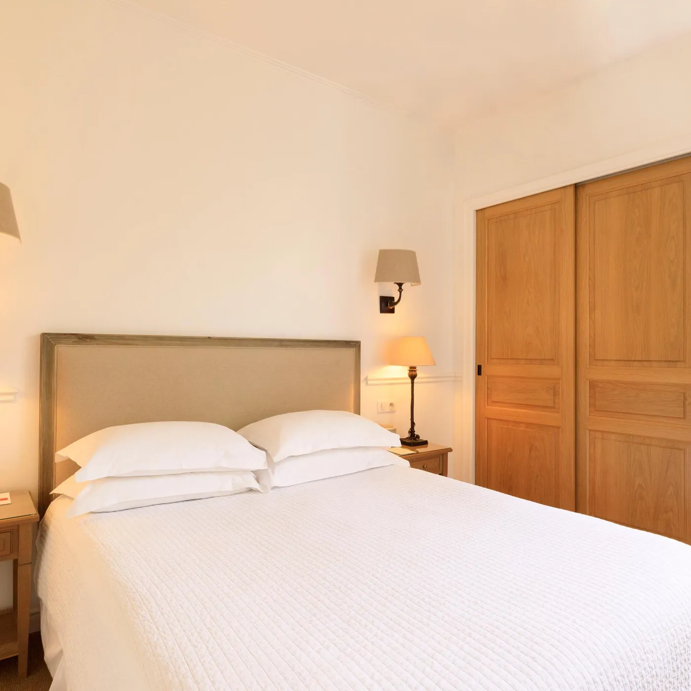 Room with a double bed, two bedside tables with lamps, a wooden door, and a window overlooking plants.