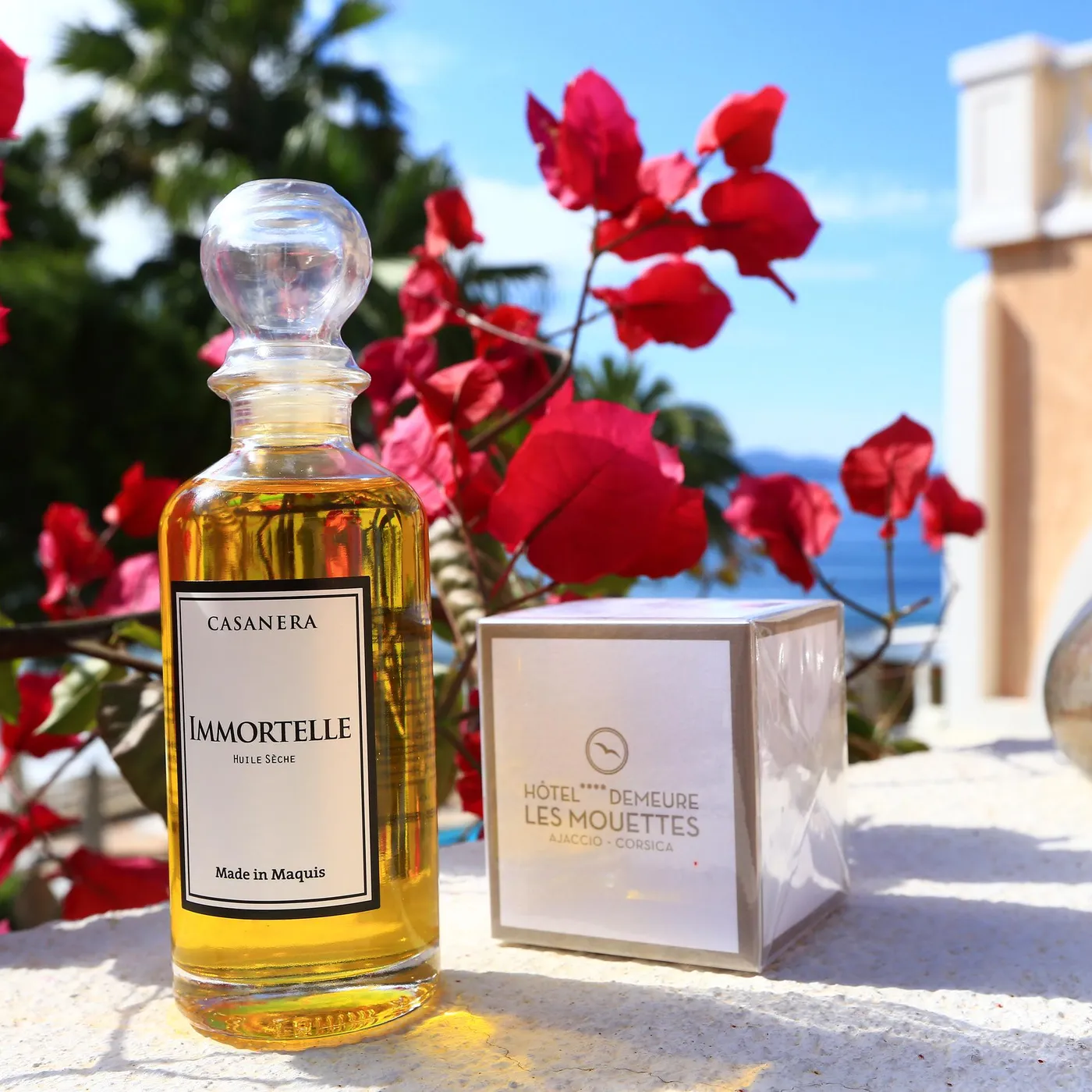 Bottle of perfume "Casanera Immortelle" and box from the hotel "Les Mouettes" placed on a stone table with red flowers in the background