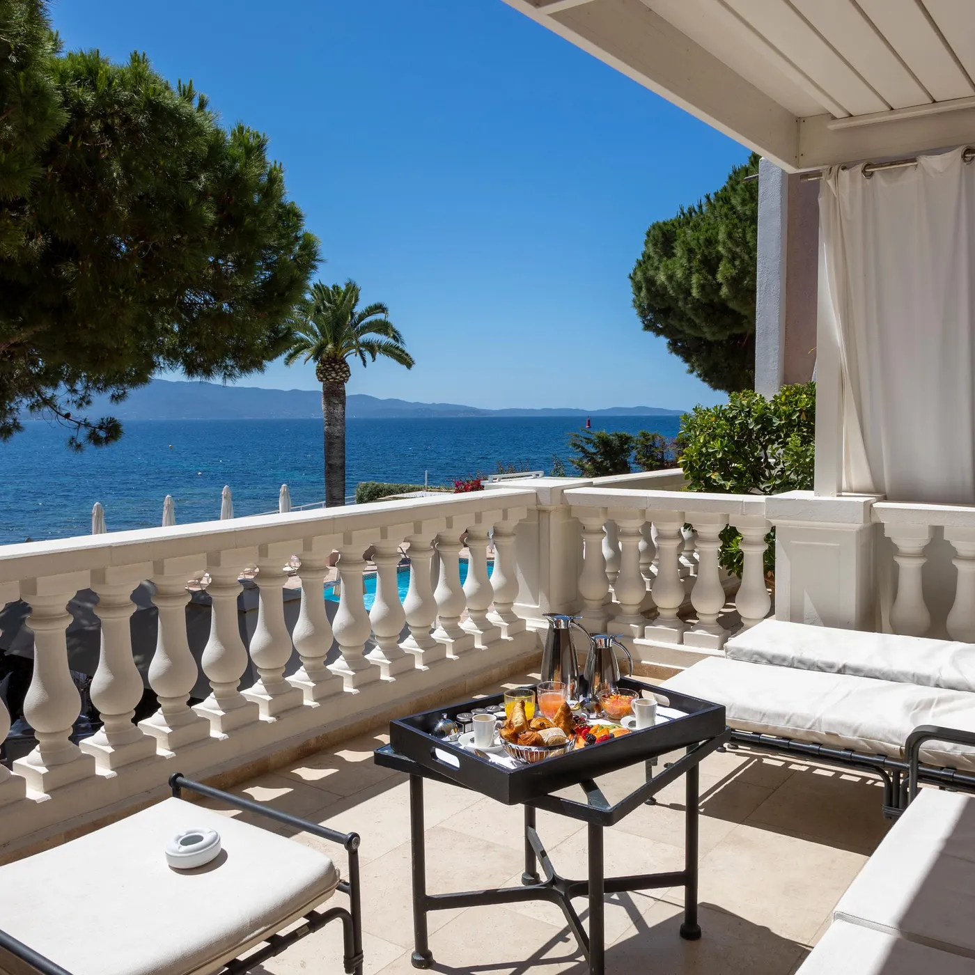 Balcon with sea view, equipped with sun loungers, a table with drinks and food, surrounded by a stone balustrade and white curtains.