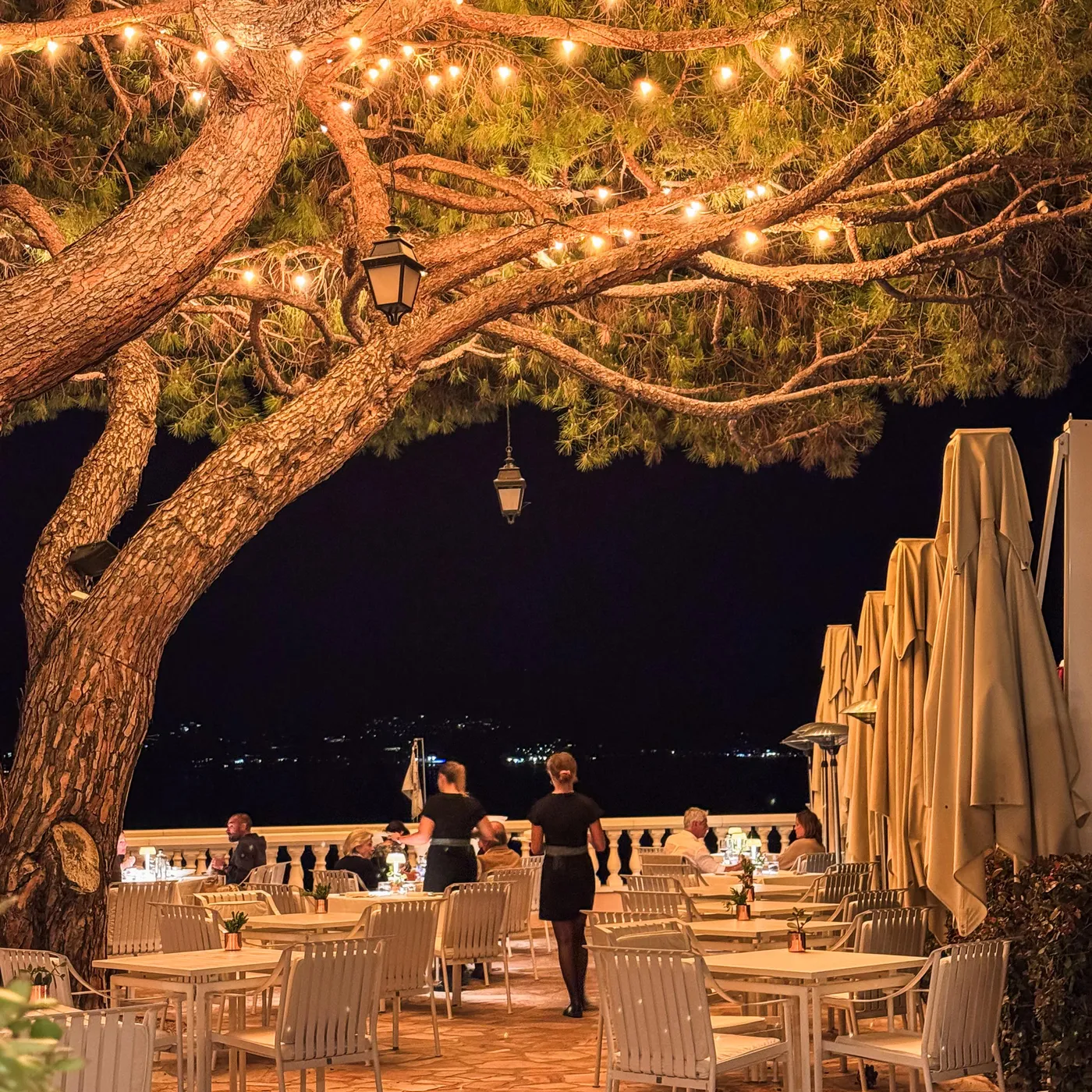 A restaurant terrace in the evening with tables and chairs, lit by string lights hanging from a tree and lanterns.