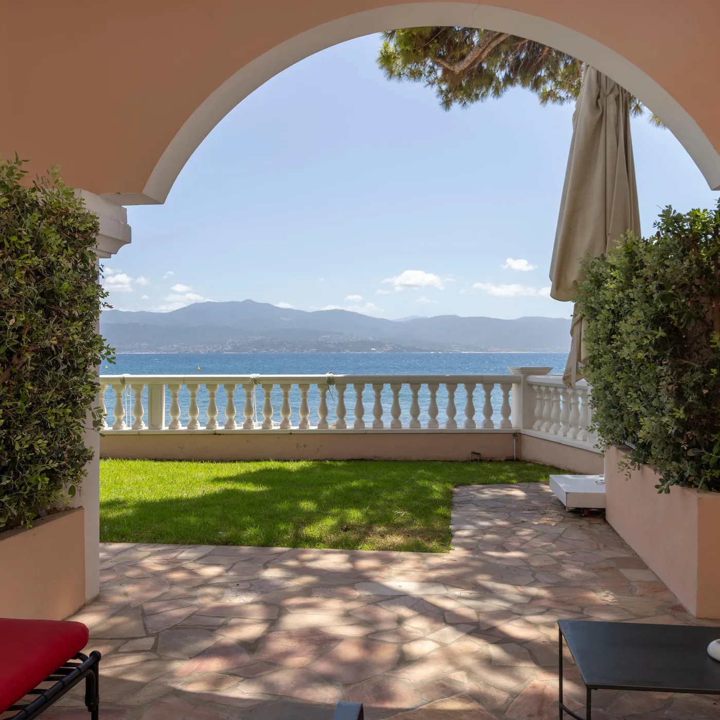 Terrace with a view of the sea and mountains, framed by arches and green plants, with a table and a chair in the foreground.