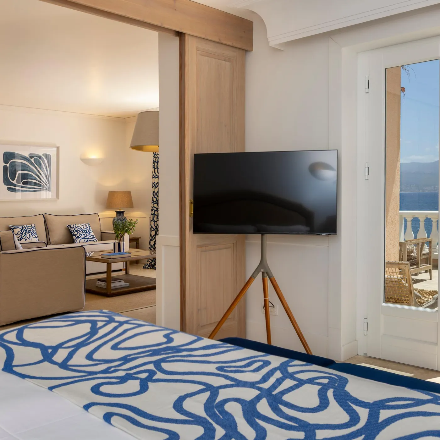 Room with blue and white patterned bed, sofa, tripod-mounted television, and view of balcony with distant sea
