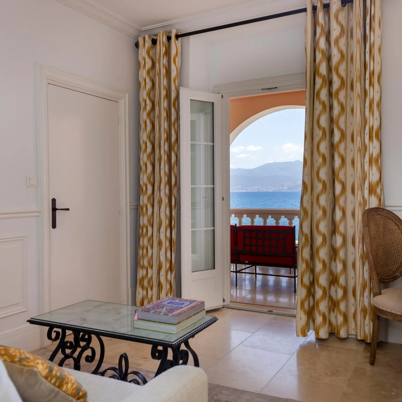 Room with sea view through an open French door, patterned curtains, coffee table with magazines, lamp and desk