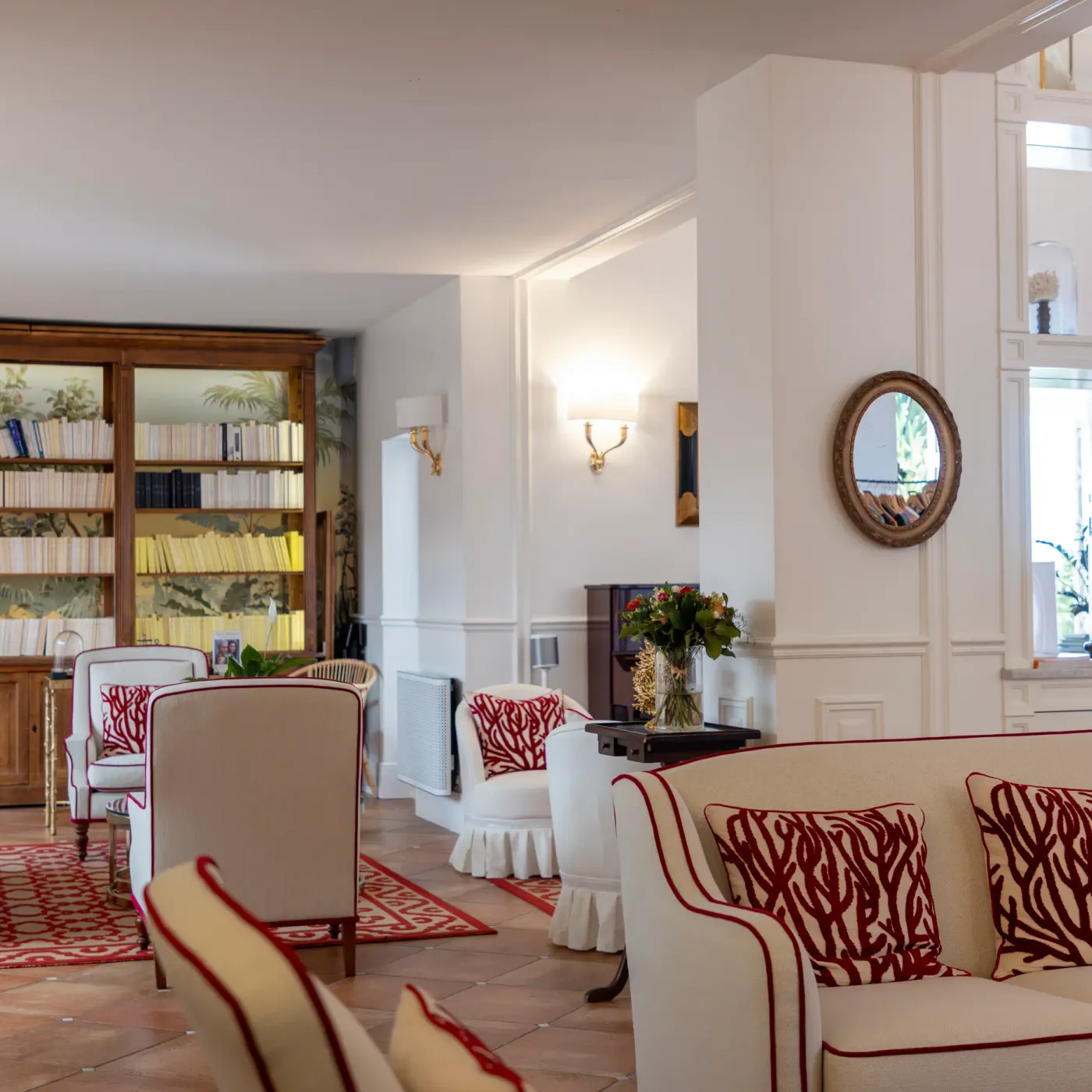 Living room with sofa and armchairs featuring red and white patterns, book-filled library, round mirror and wall decorations.