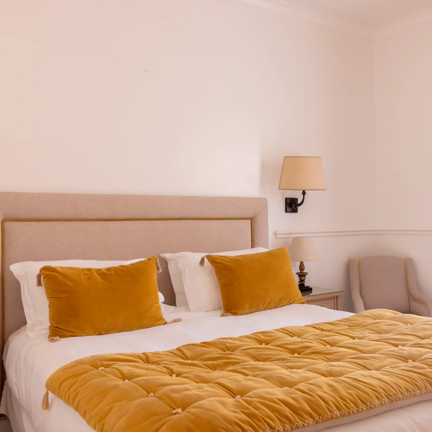 Double bed with a beige headboard, a blanket and gold cushions, two bedside tables with lamps, and patterned curtains.