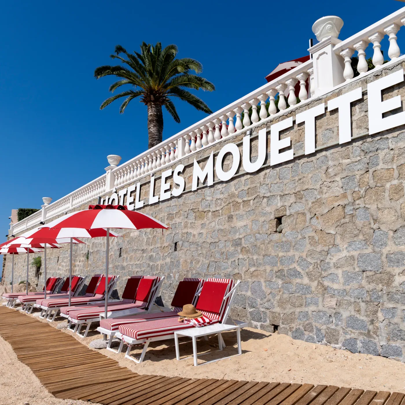 Une rangée de transats rouges et blancs sous des parasols rouges et blancs devant un mur de pierre avec l'inscription "HOTEL LES MOUETTES" et une balustrade blanche, avec une vue sur la mer au loin