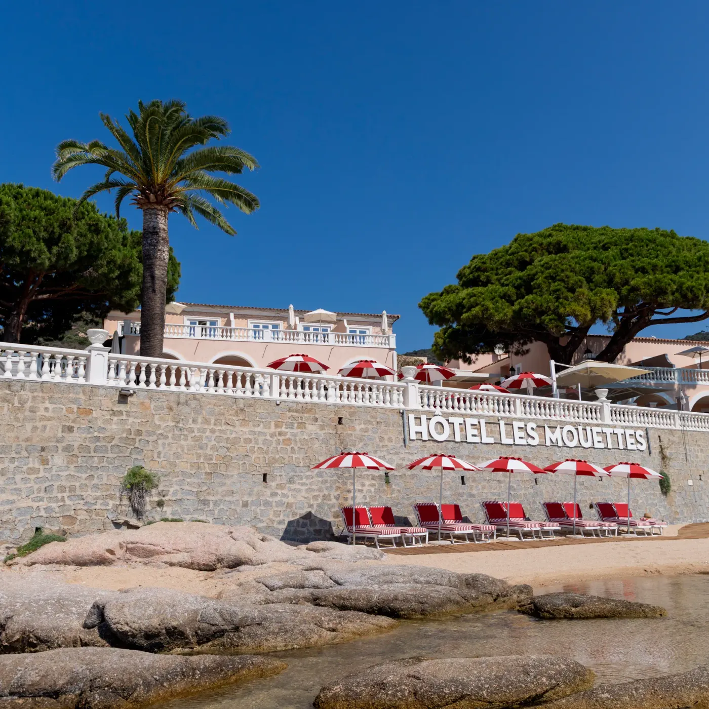 Vue de l'hôtel Les Mouettes avec une plage de galets, des transats et des parasols rouges
