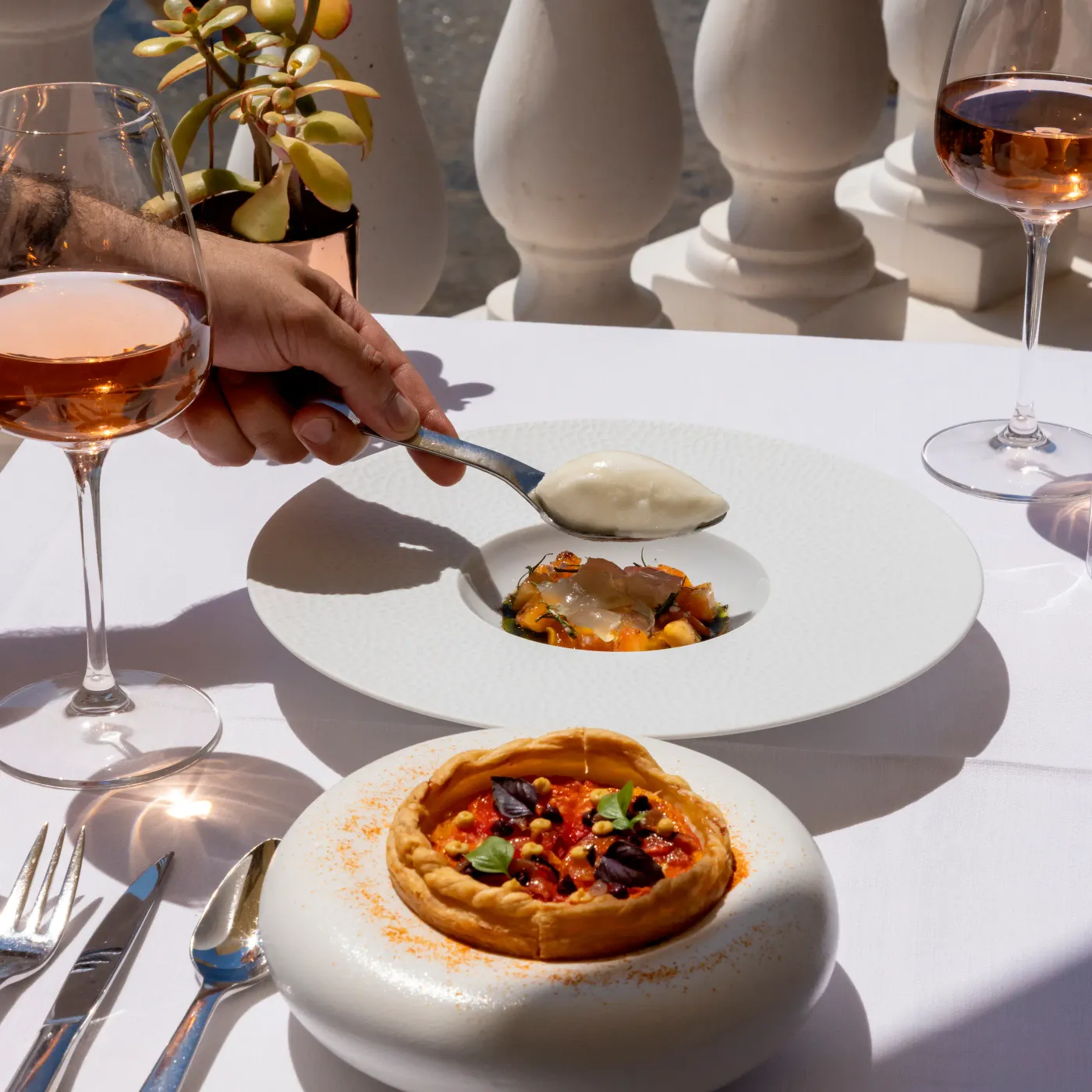 A person holds a spoon above a plate with a small portion of food, accompanied by wine glasses and an individual tart on a white table.
