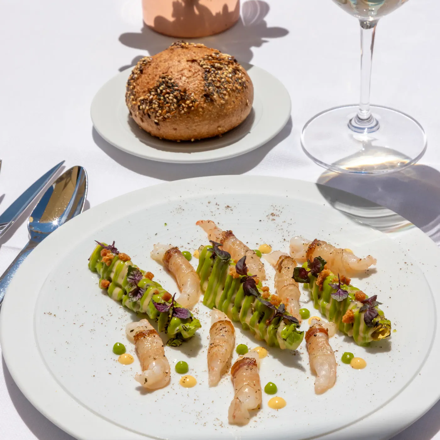 Plate with green asparagus wrapped in ham slices, served with peas and sauce, a small bread roll, and a glass of white wine beside it.
