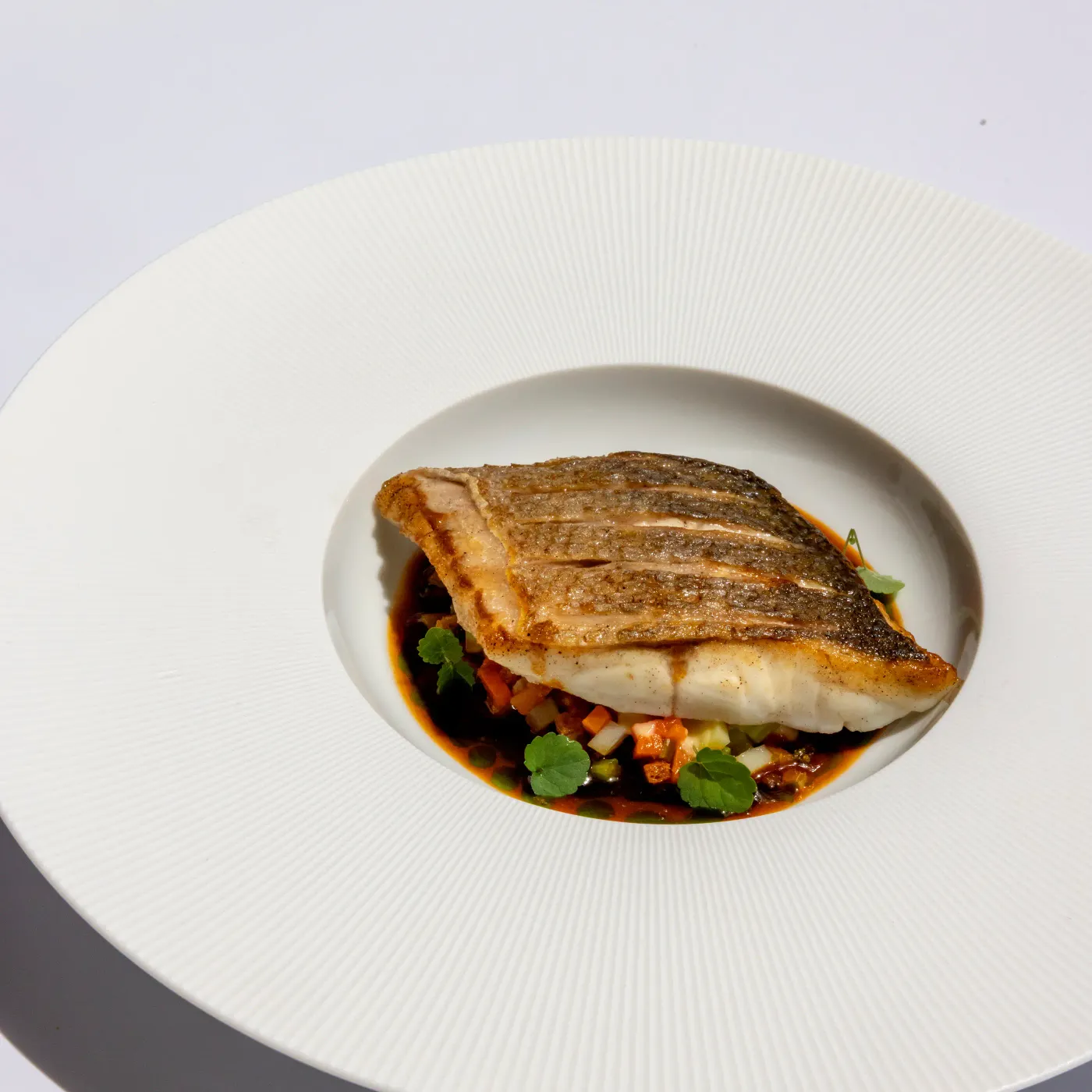 A grilled fish fillet on a bed of vegetables and sauce in a white dish