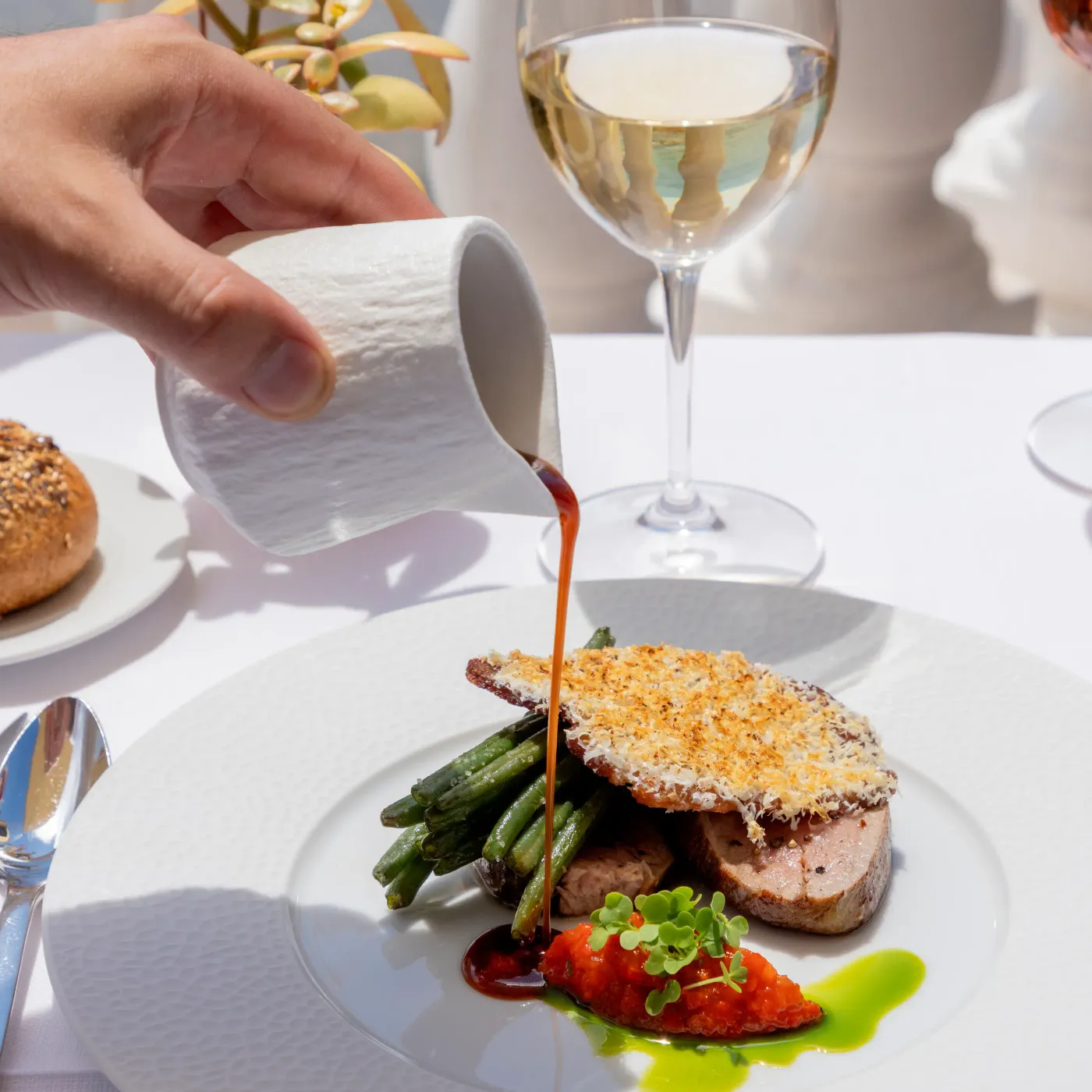 A hand pours a sauce over a plate containing a dish made of meat, asparagus, and a vegetable garnish, accompanied by a glass of white wine.
