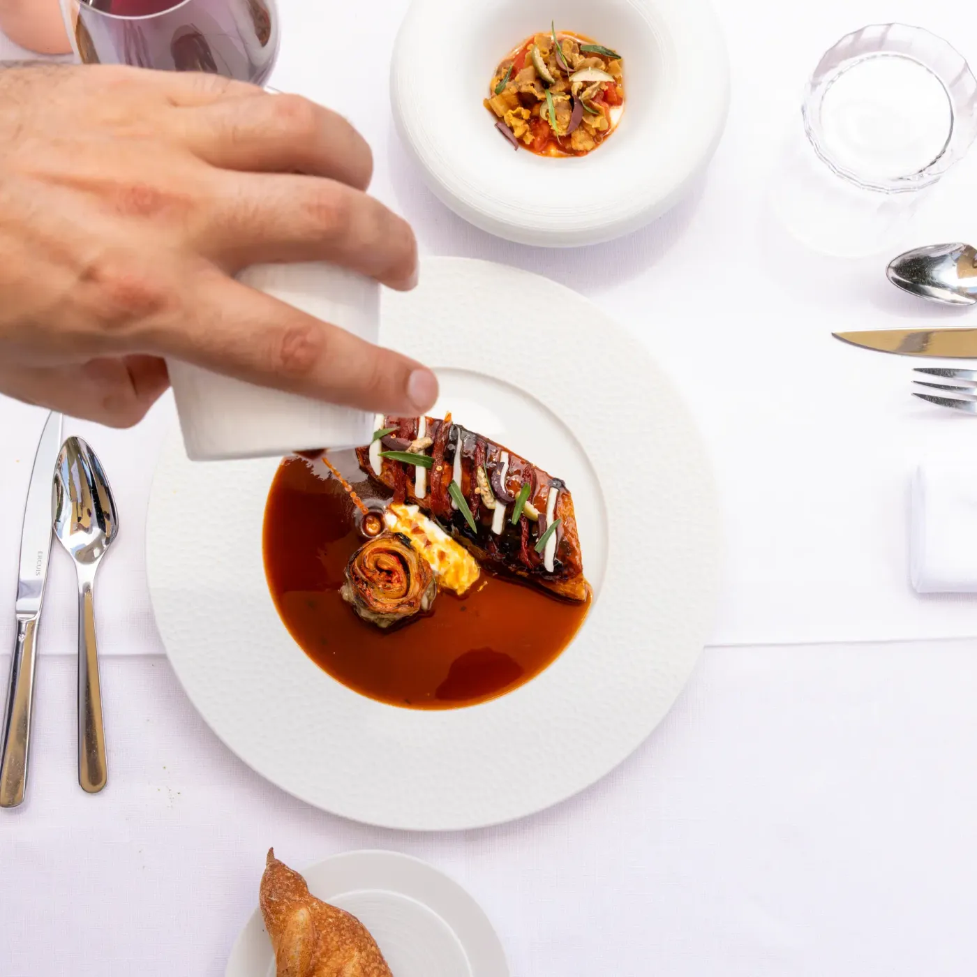 A hand holding a napkin approaches a plate containing a dish with sauce and garnishes, on a table set with cutlery and glasses.