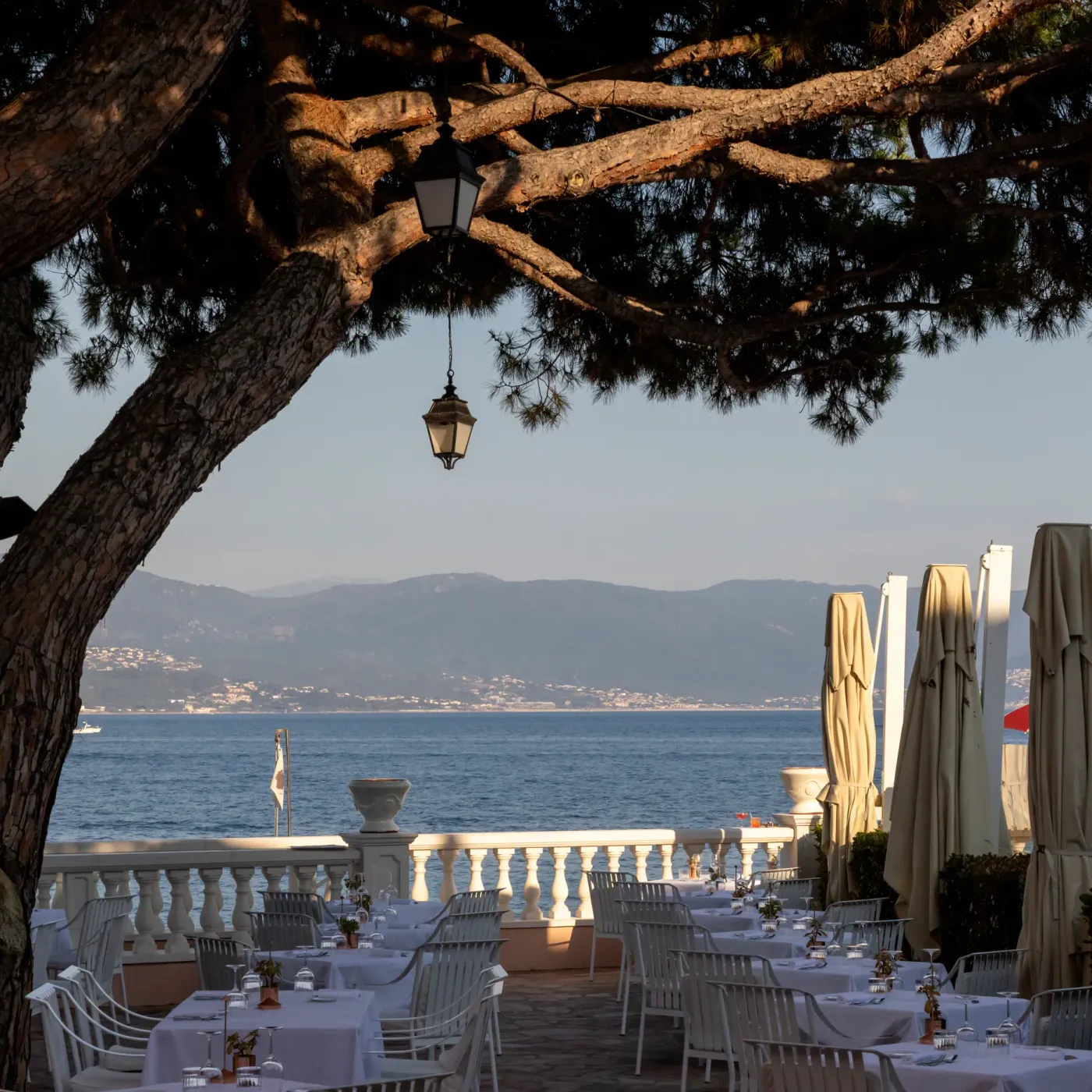 Terrasse de restaurant en bord de mer avec tables et chaises, vue sur la mer et les montagnes au loin