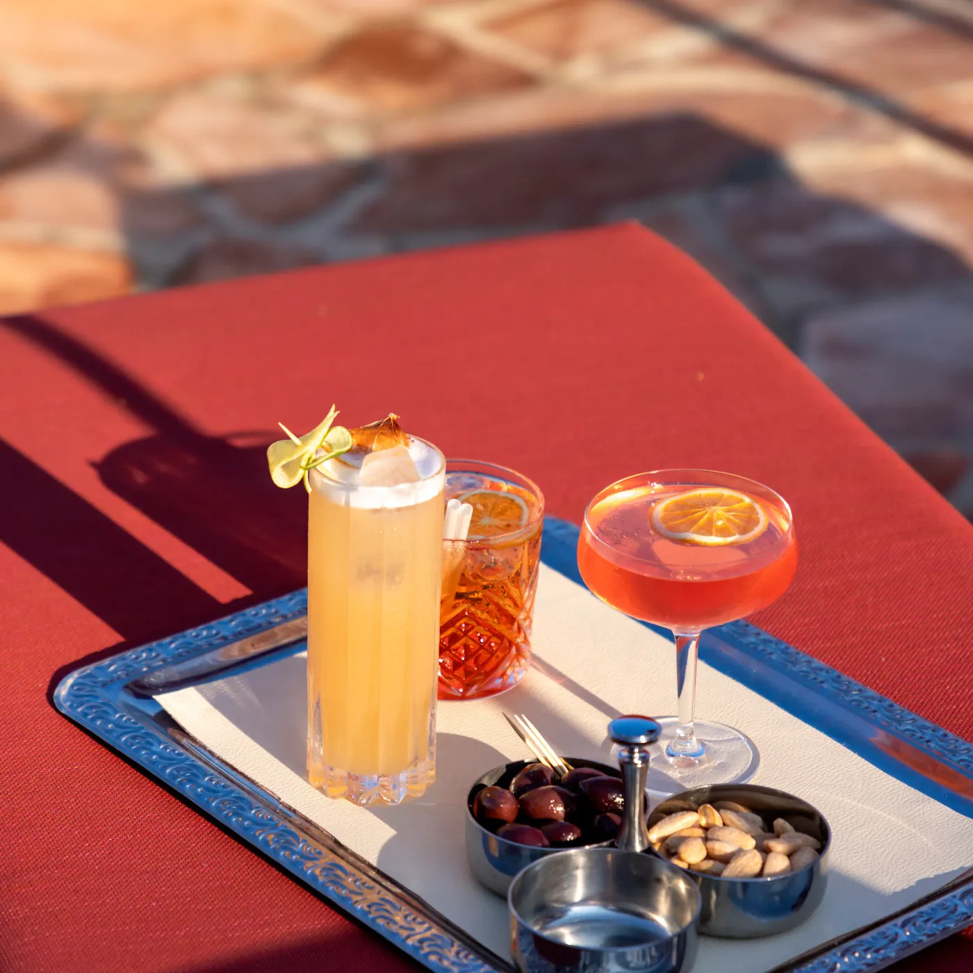 A tray with three glasses of different drinks and small bowls of nuts and olives on a red table