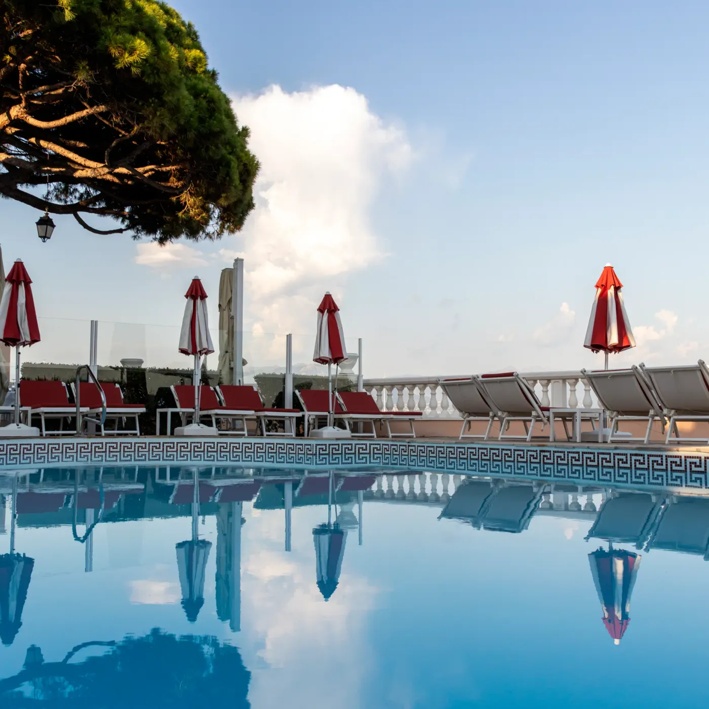 Piscine avec des transats et des parasols rouges et blancs, reflétant le ciel et les arbres dans l'eau calme