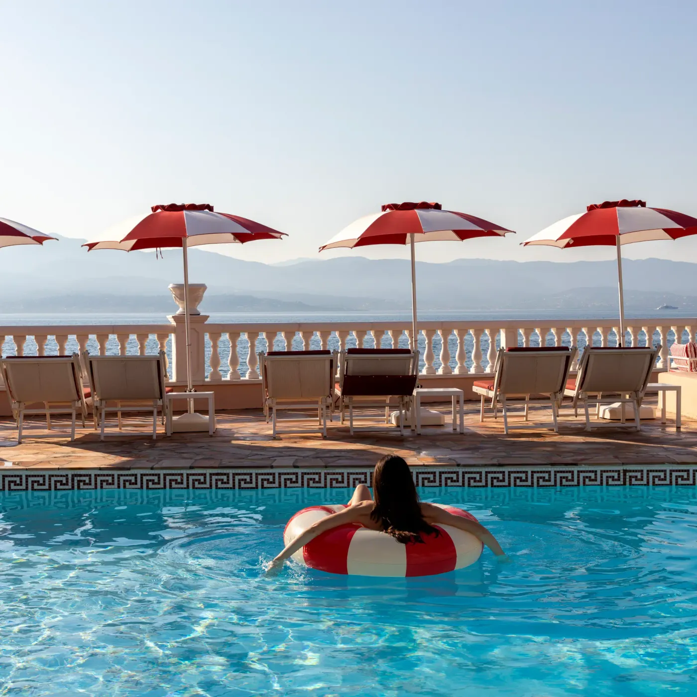 Une personne sur un bouée dans une piscine avec vue sur la mer et des transats à l'ombre de parasols