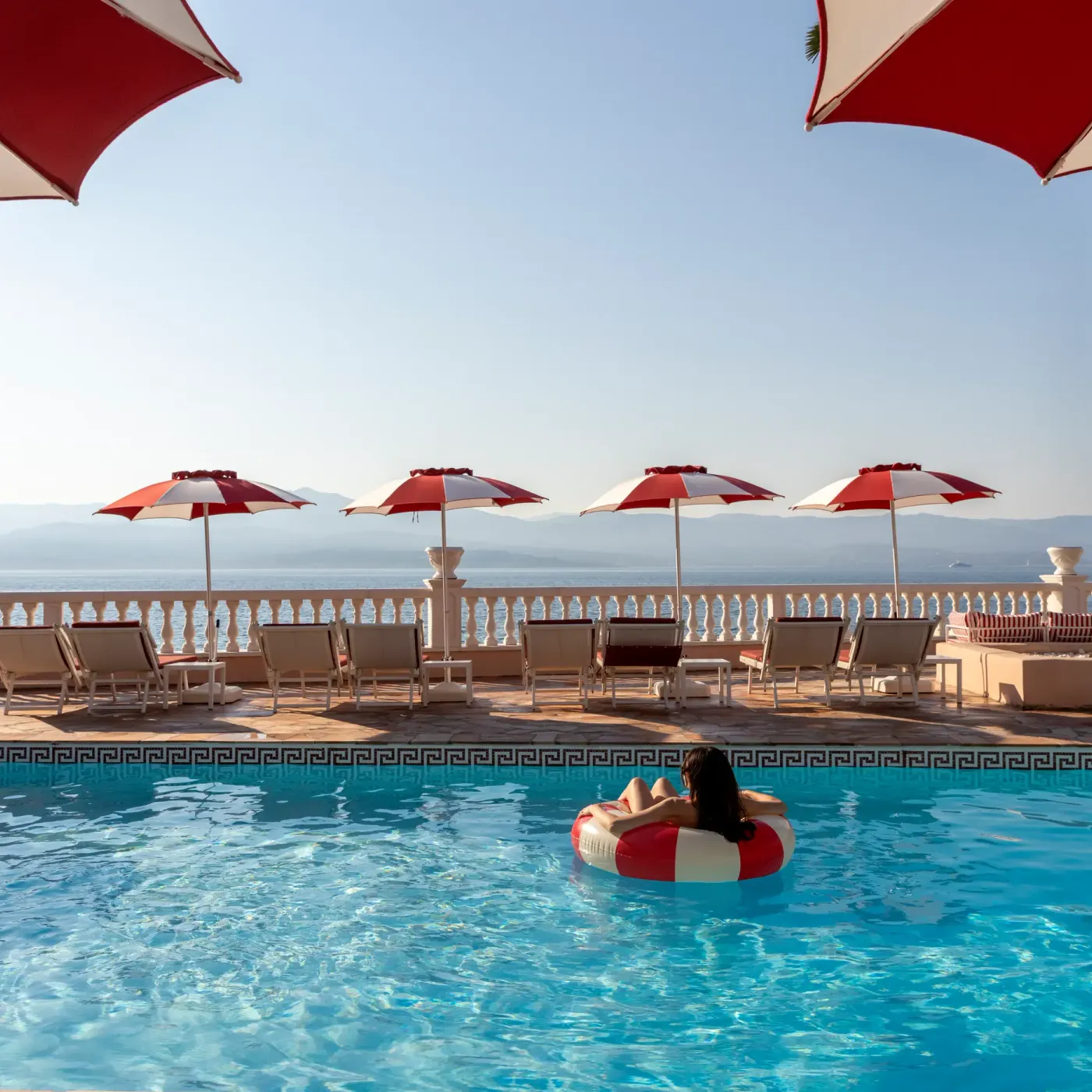 Une personne sur un bouée en forme de donut dans une piscine avec vue sur la mer et des chaises longues à l'ombre de parasols