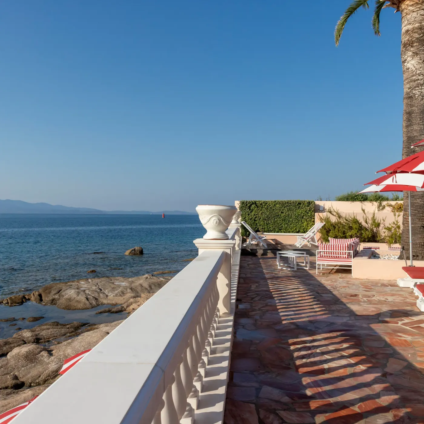 Une terrasse en bord de mer avec des transats, des parasols et une balustrade blanche, face à une étendue d'eau calme et des montagnes au loin