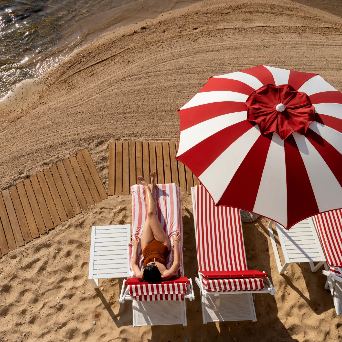 Une personne allongée sur un transat à côté de la mer, sous un parasol rayé rouge et blanc