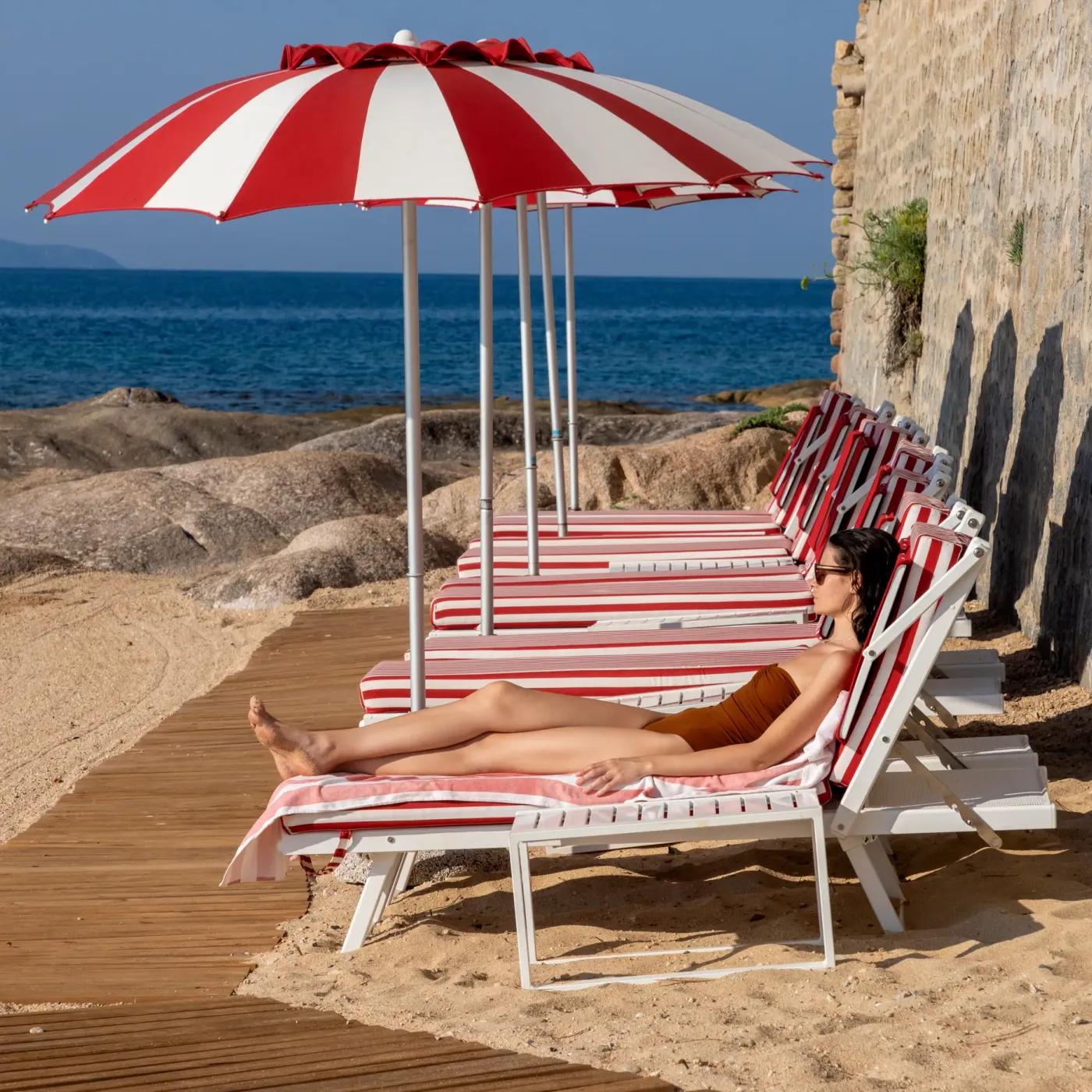 Une personne en maillot de bain bronze sur une chaise longue sous un parasol à rayures rouges et blanches, face à la mer