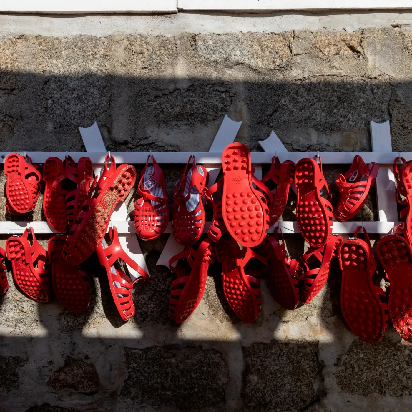 Des chaussures rouges en plastique sont accrochées sur une barre métallique fixée à un mur en pierre