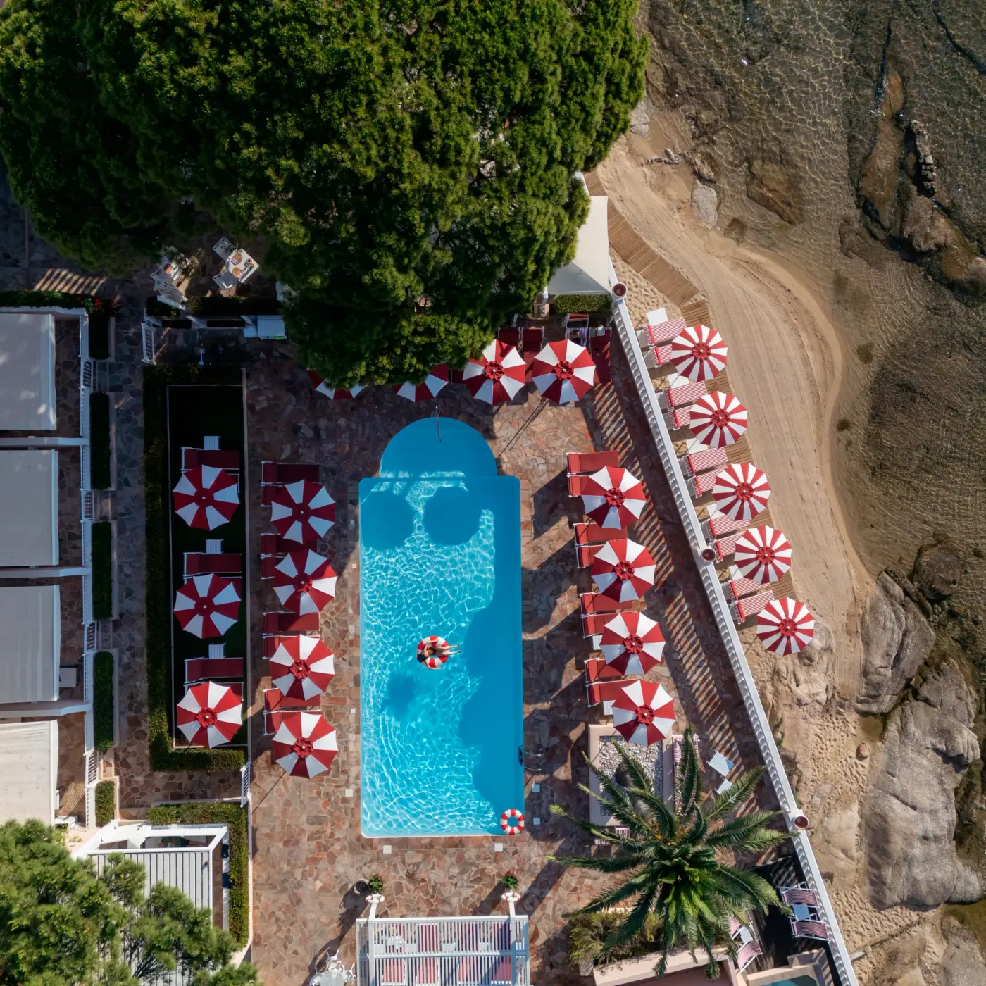 Vue aérienne d'une piscine rectangulaire entourée de transats et de parasols rouges et blancs, située près d'un bâtiment et d'une zone rocheuse