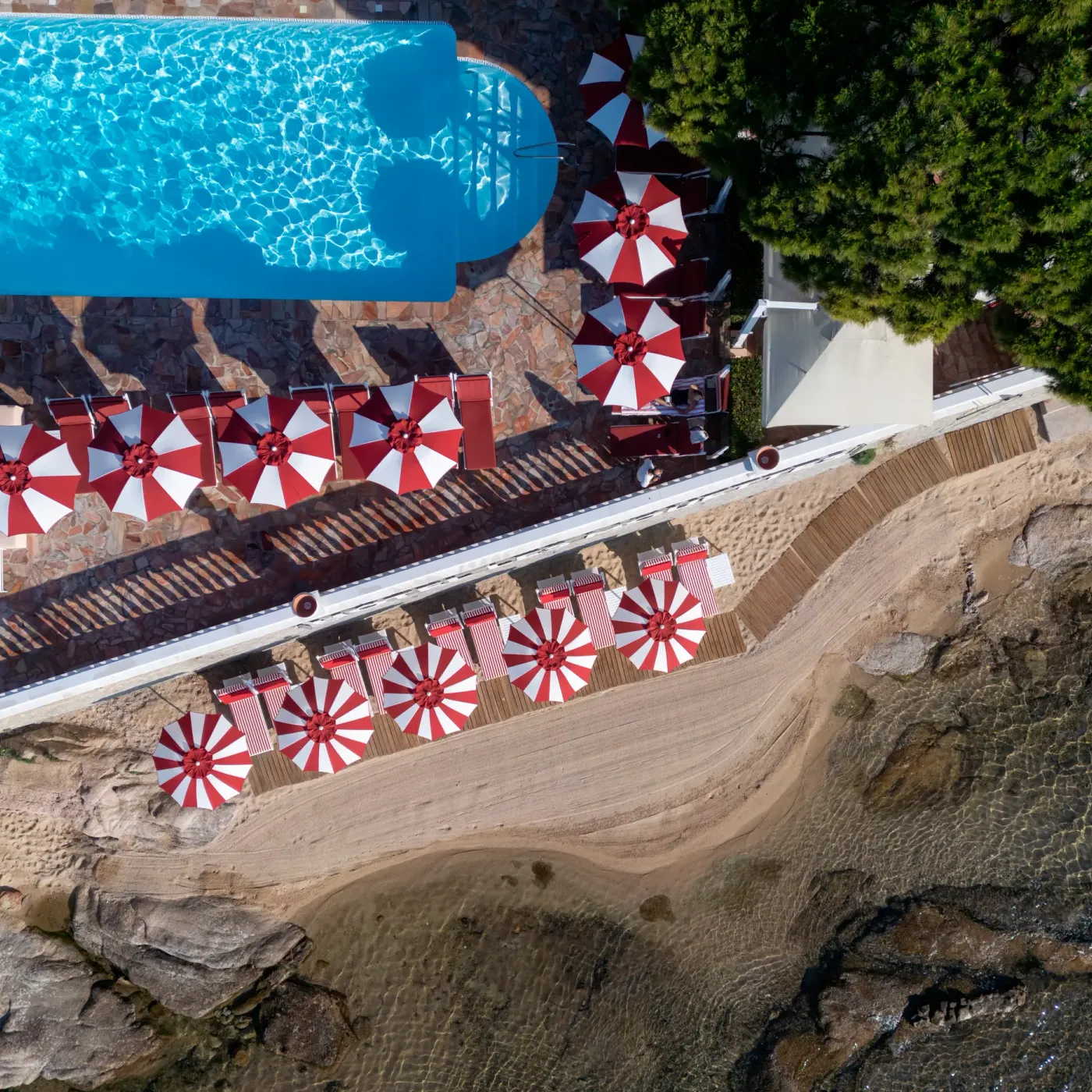 Vue aérienne d'une piscine avec des parasols rouges et blancs et des transats sur une plage rocheuse