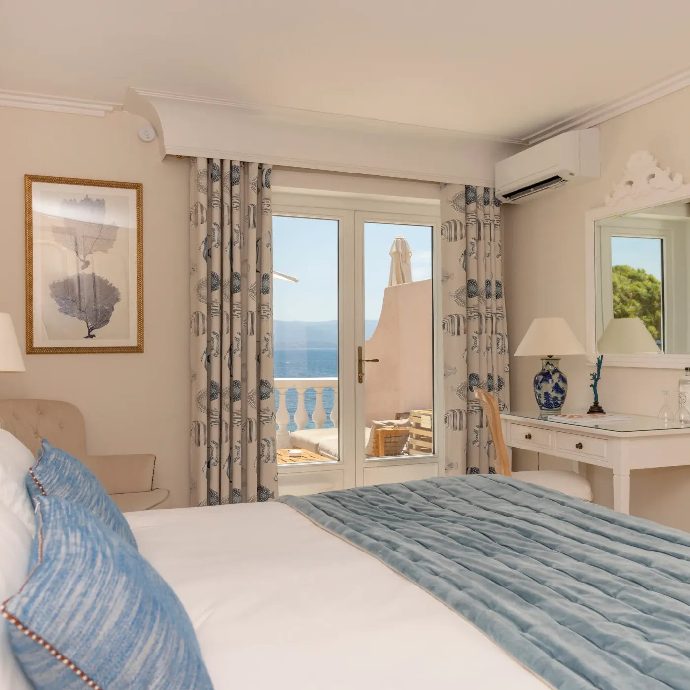Room with a bed covered by a blue blanket, a French window offering a sea view, a mirror, a television, and a bedside lamp.