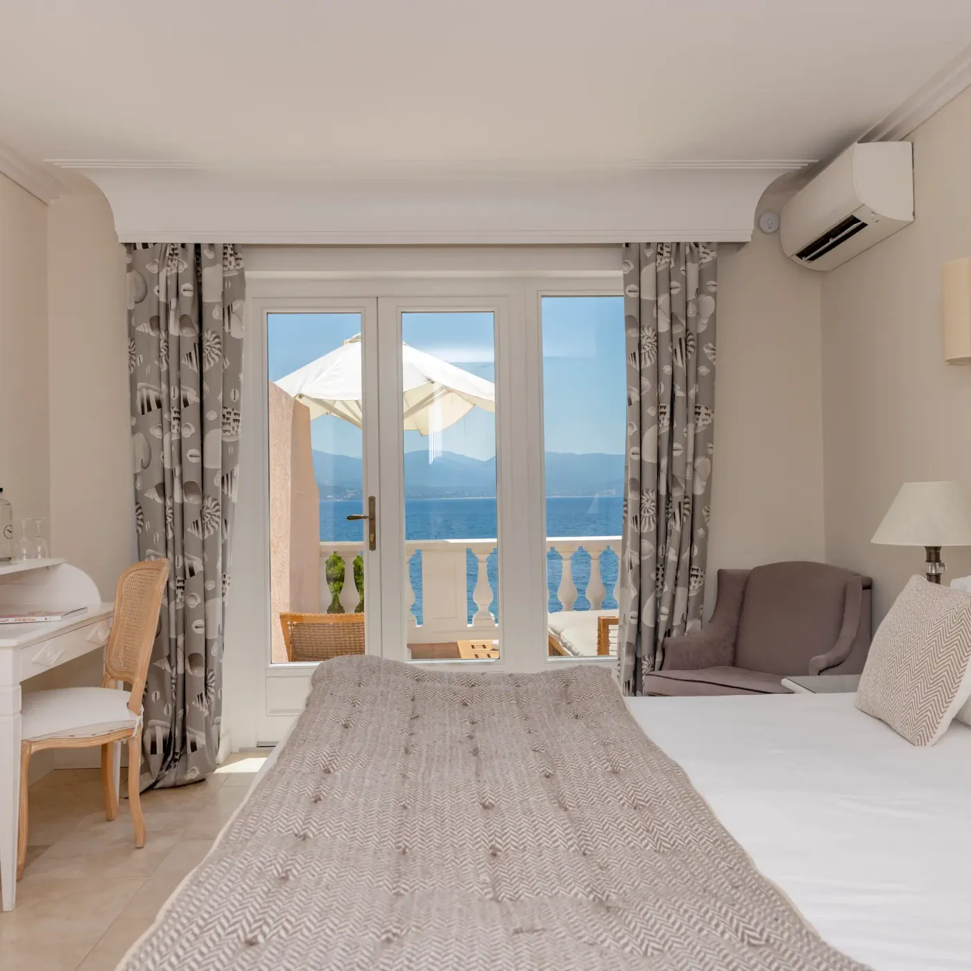 Room with double bed, desk, French door opening onto a balcony with sea and mountain views, grey curtains and soft lighting