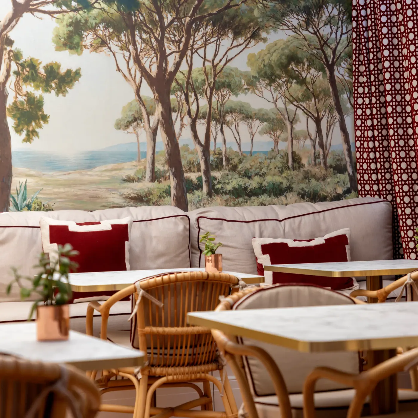 An outdoor space with tables, wicker chairs, and a white sofa with red cushions, in front of a painted wall depicting a forest and a coastal landscape.