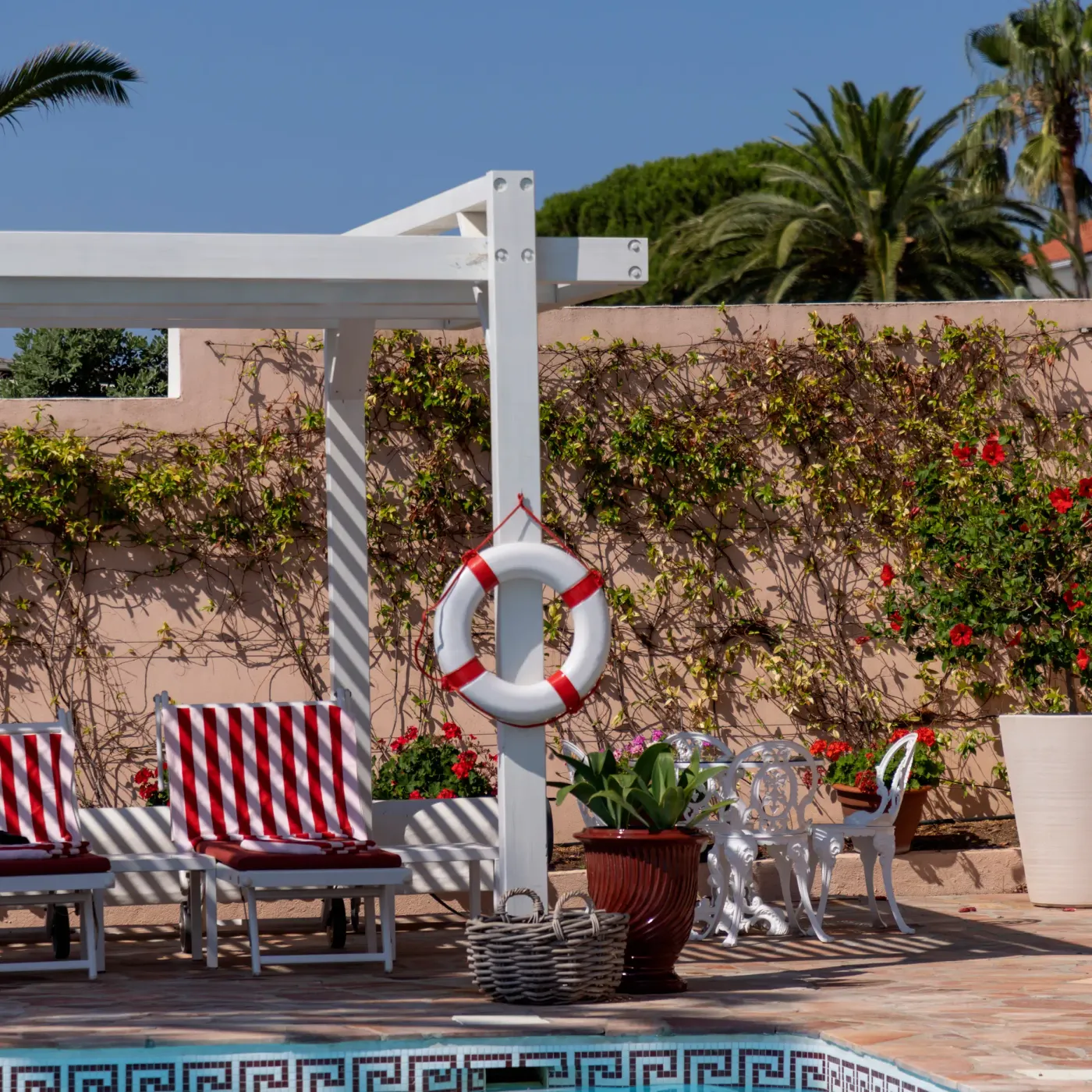Patio avec transats rayés rouges et blancs, chaises blanches, pots de fleurs et une vieillesse de sauvetage accrochée à un poteau près d'une piscine