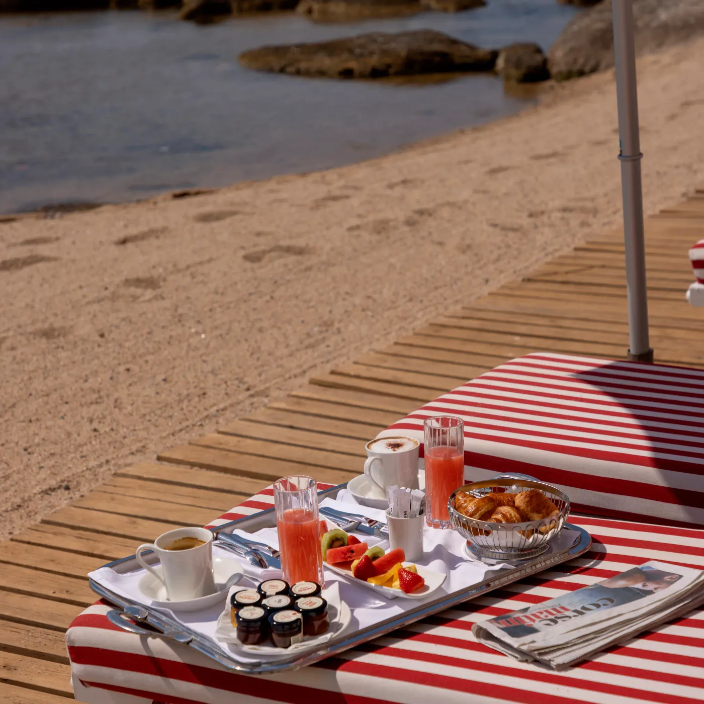 Un plateau-repas avec café, jus d'orange, croissants, fruits et confitures posé sur une table en bois près d'une plage