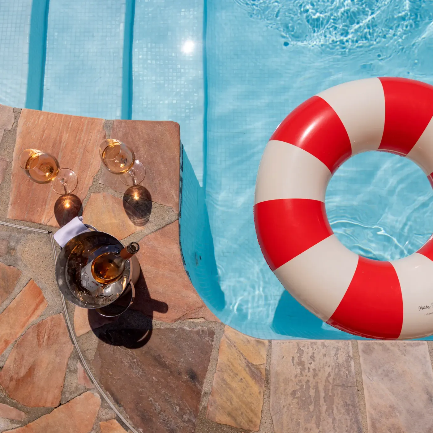 Un verre de cocktail, une bouteille et des accessoires sont posés sur le bord d'une piscine à côté d'un bouée de sauvetage rouge et blanc