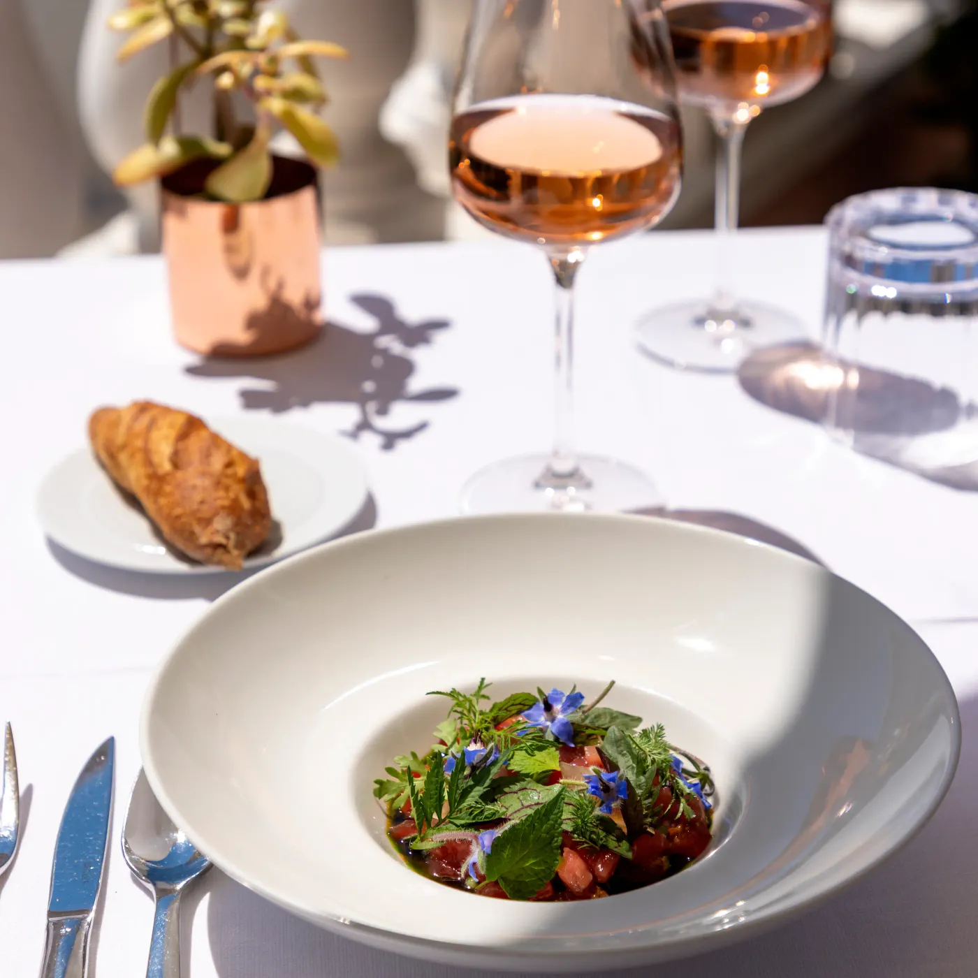 A table set with a salad dish, wine glasses, a baguette, and a small potted plant.
