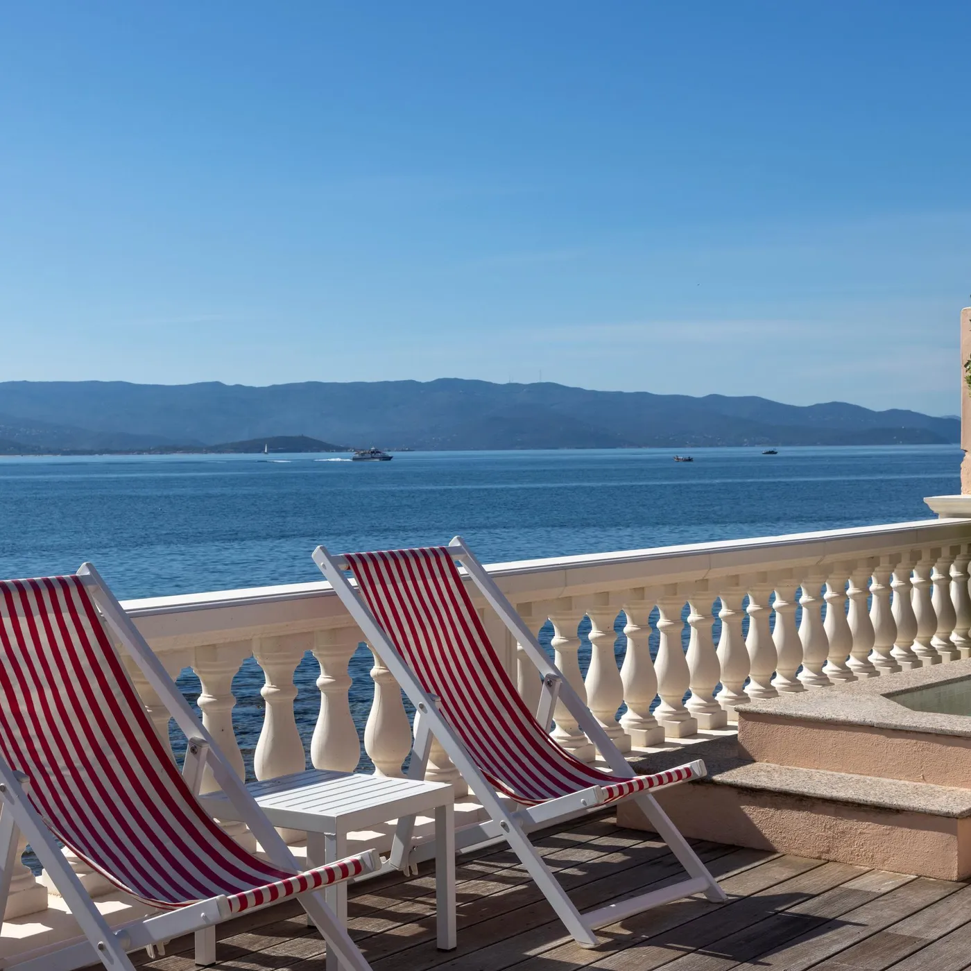 Deux transats à rayures rouges et blanches sont placés sur une terrasse en bois face à une mer calme, avec des montagnes en arrière-plan