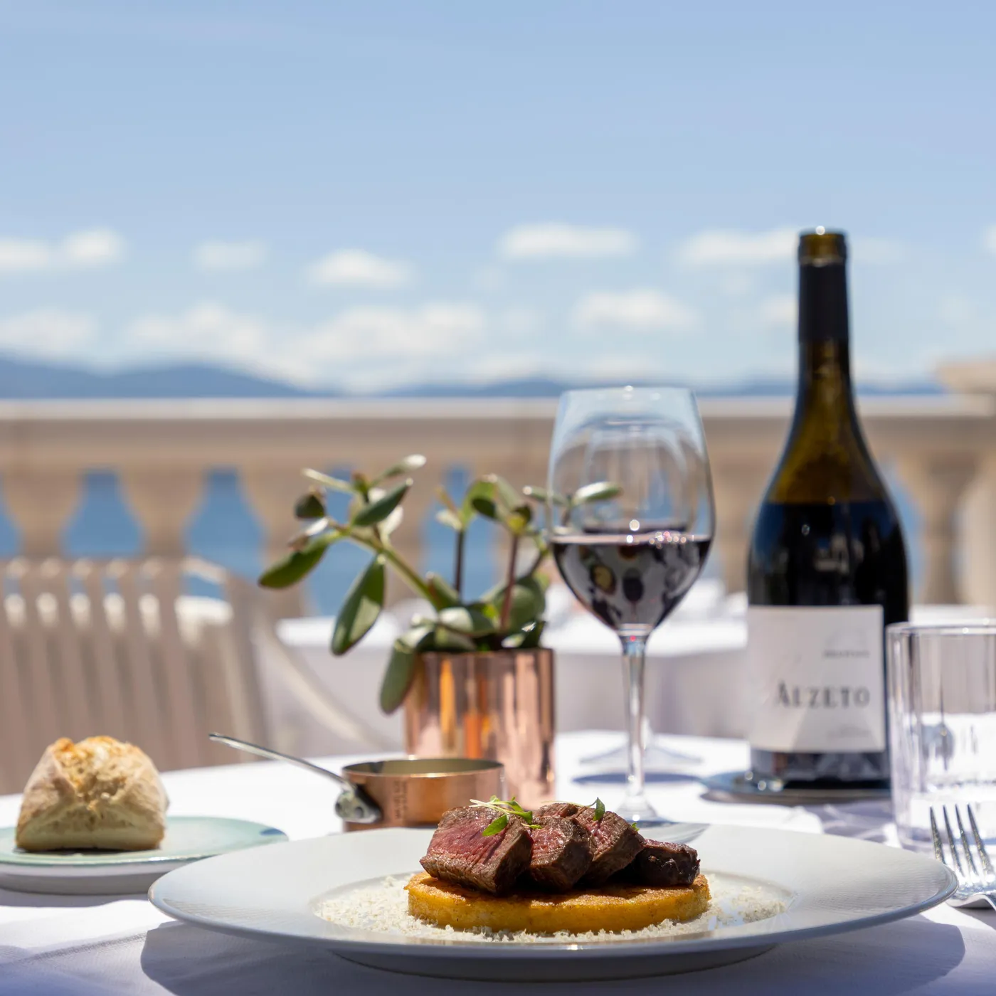 A plate of grilled meat with polenta, a glass of wine, and a bottle on a terrace table with a sea view.