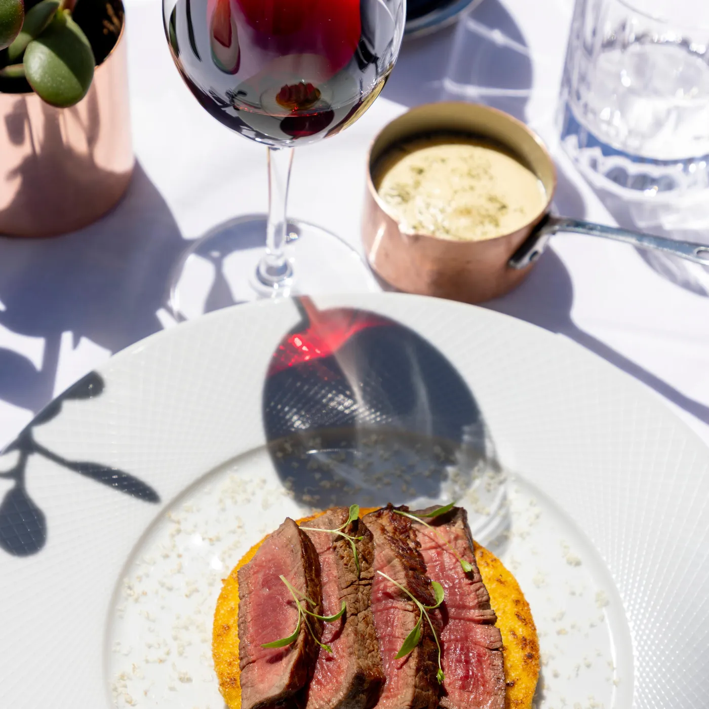A glass of red wine, a plate with slices of meat on a flatbread, and a bowl of sauce on a white table