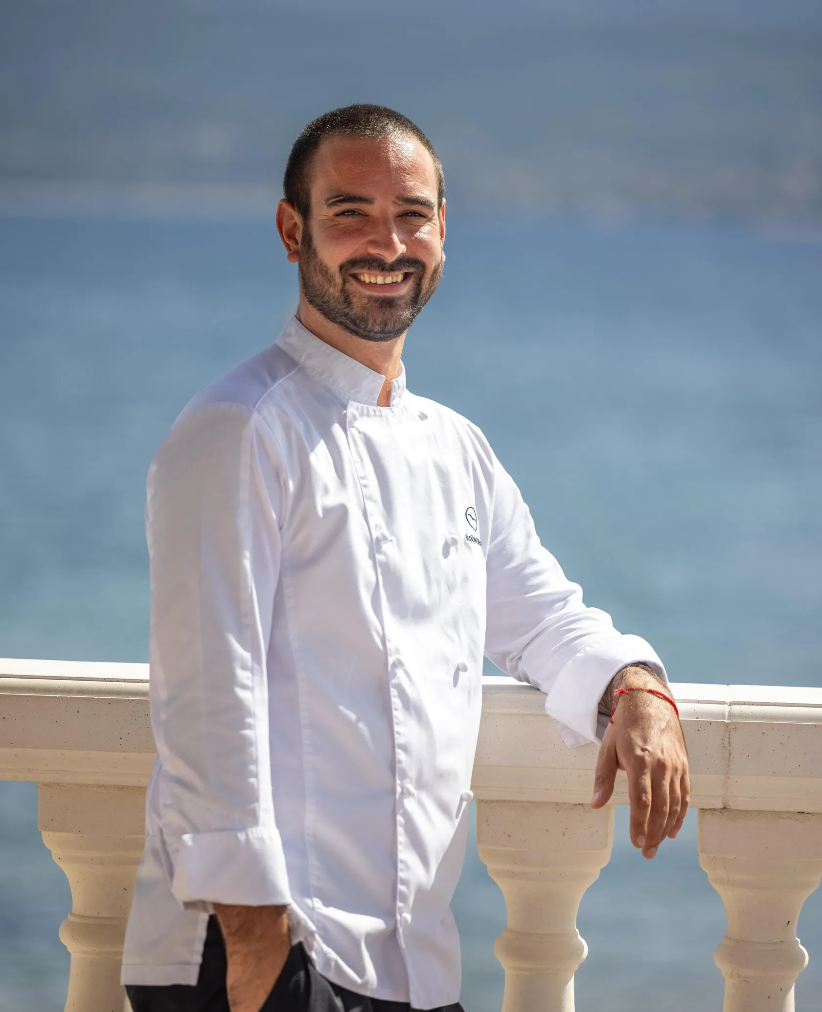 Un homme en chemise blanche de chef pose en souriant, appuyé sur une balustrade avec le ciel bleu en arrière-plan