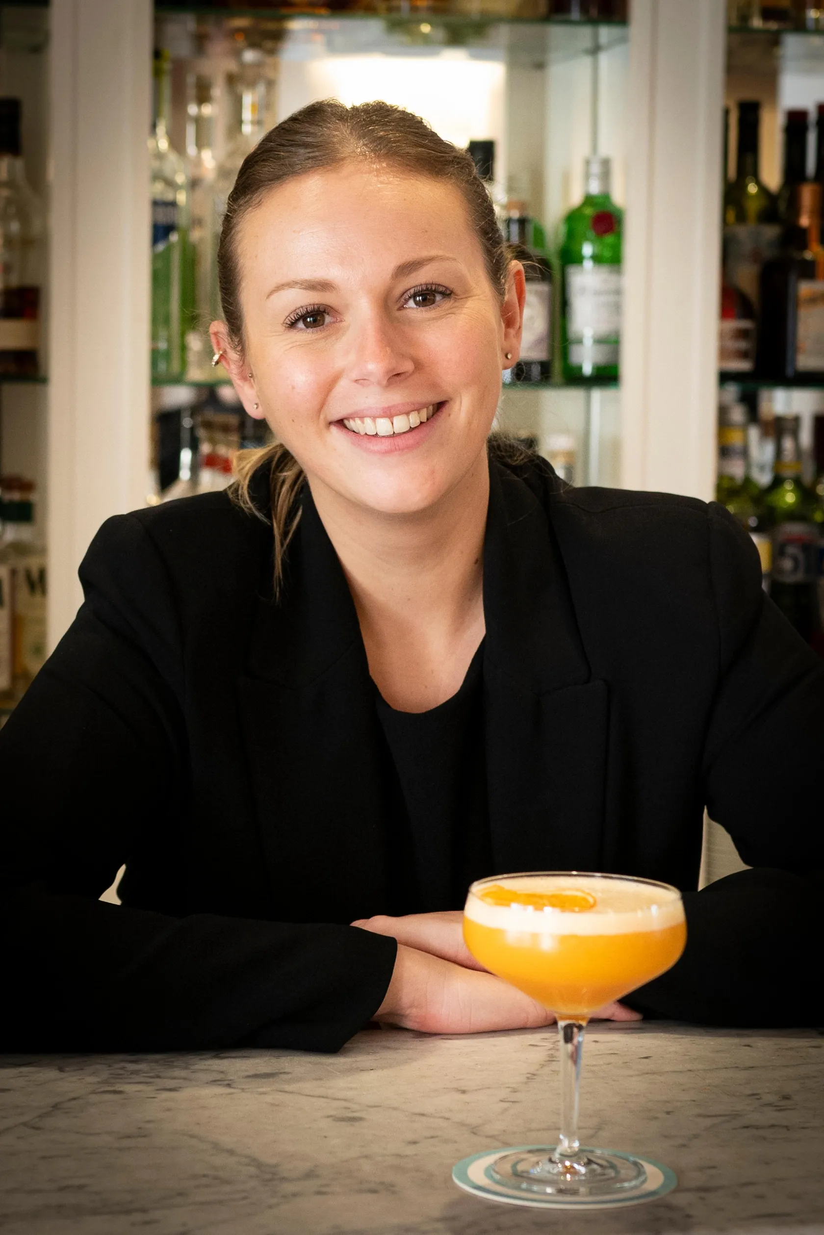 Une femme souriante en tenue noire derrière un bar tient un verre contenant un cocktail orange