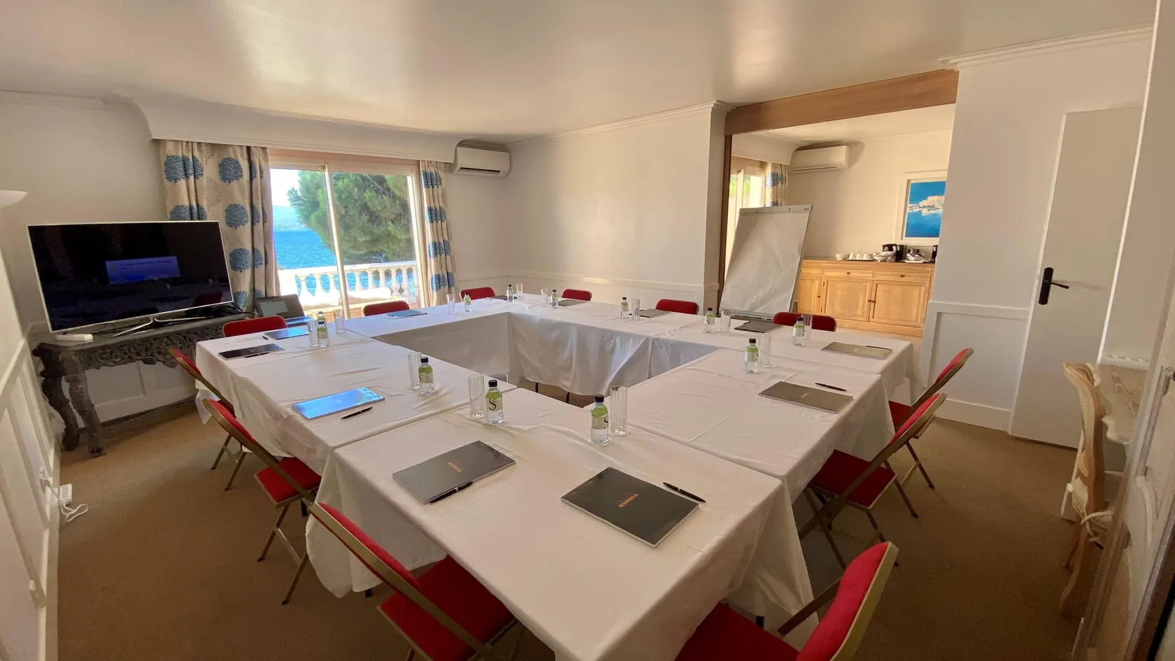 Meeting room with tables covered in white tablecloths, red chairs, television screen, and sea view through a window.