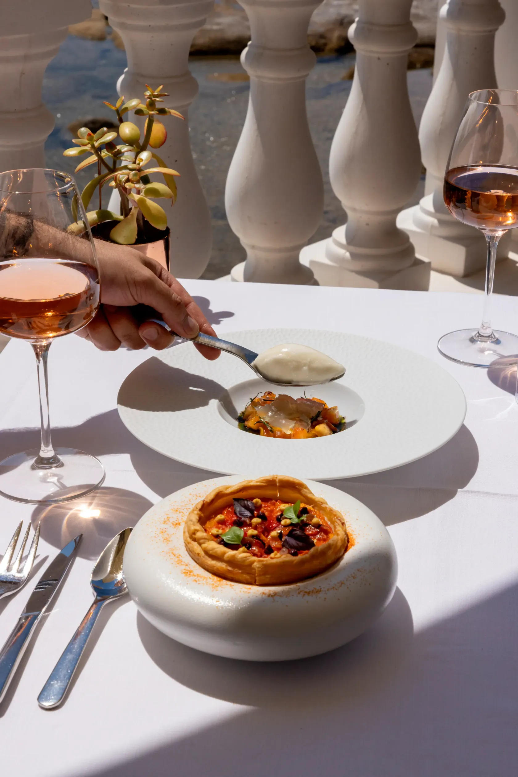 Une personne tient une cuillère au-dessus d'une assiette avec une petite portion de nourriture, accompagnée de verres de vin et d'une tarte individuelle sur une table blanche