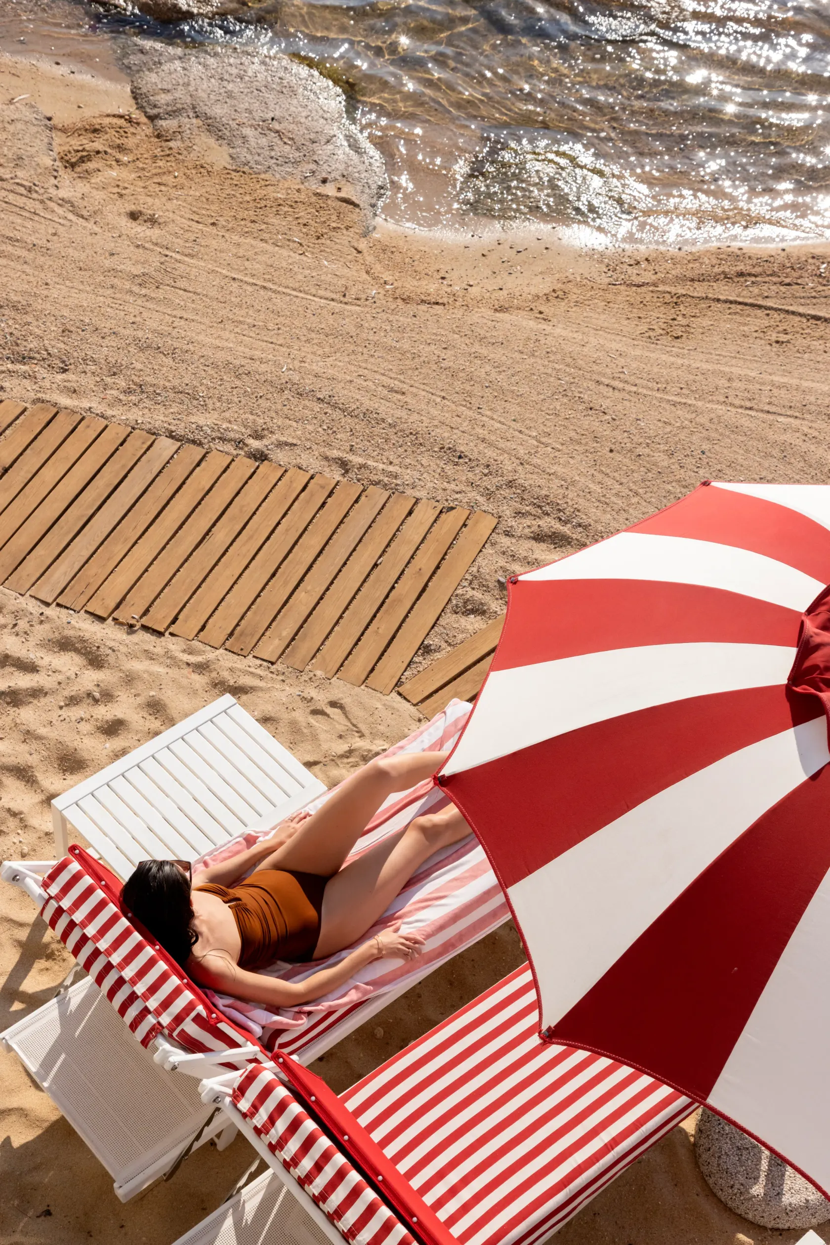 Une personne allongée sur une chaise longue à rayures rouges et blanches près de la mer, à l'ombre d'un parasol similaire