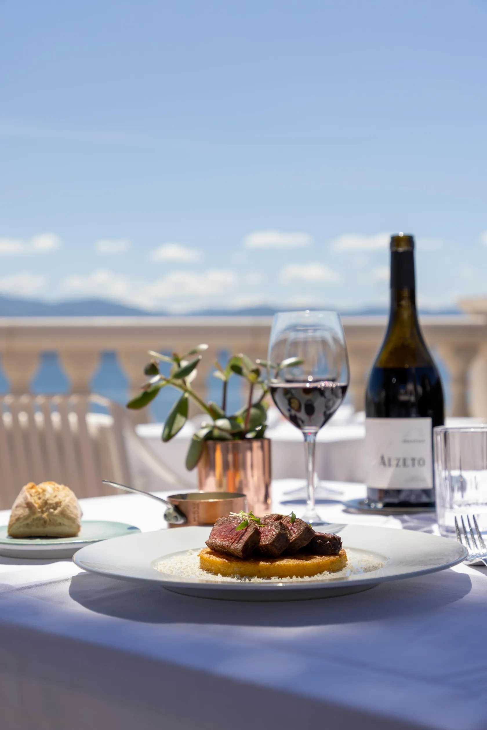 Une assiette de viande grillée avec polenta, un verre de vin et une bouteille sur une table en terrasse avec vue sur la mer