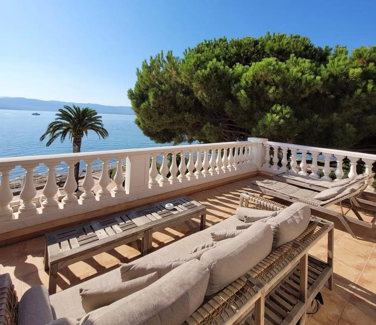 Terrace with outdoor furniture and sea view, bordered by a white balustrade and trees.