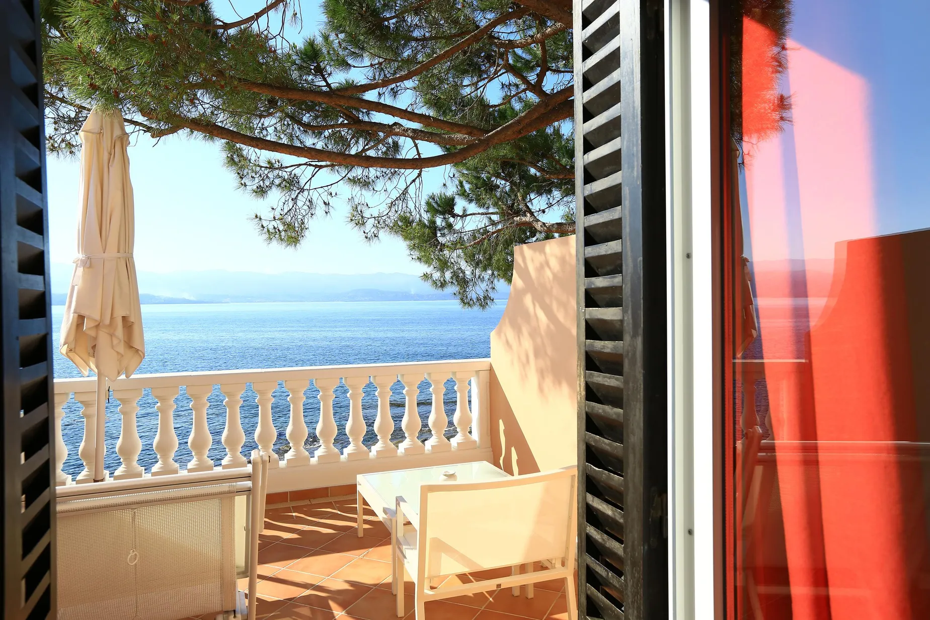 Balkon mit Meerblick, Tisch, Stühle und Sonnenschirm, umgeben von Ästen und roten Fensterläden